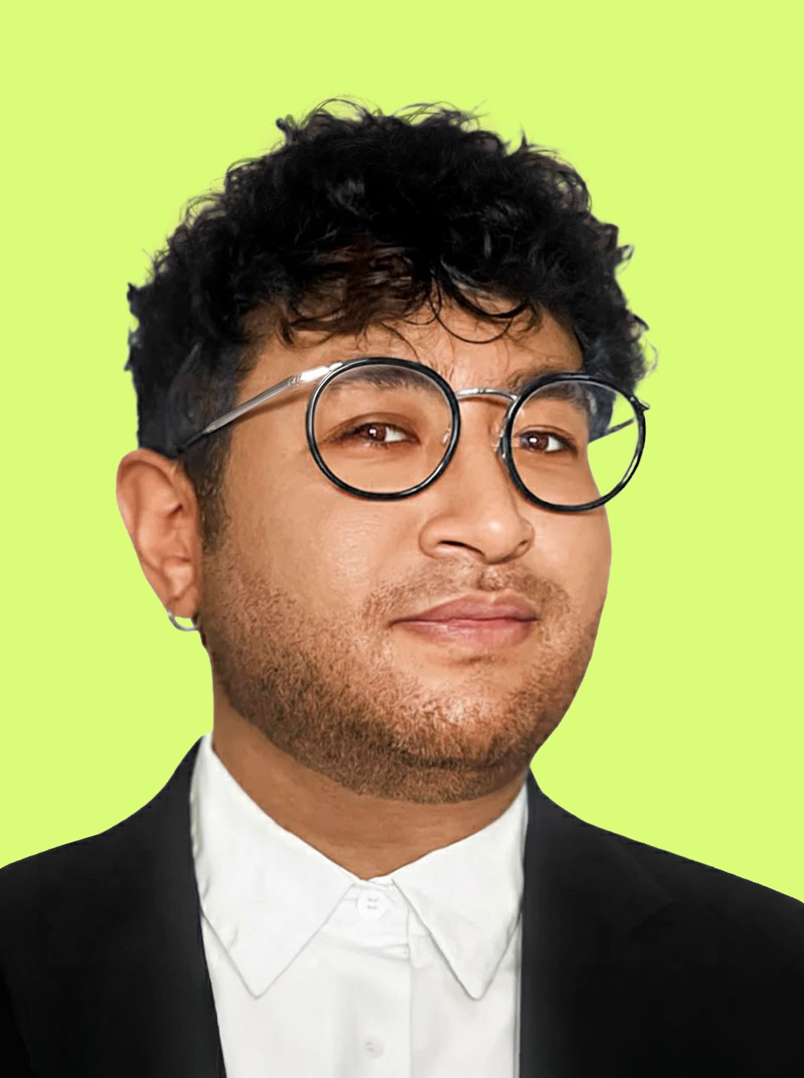 A person with short curly dark hair, glasses, and an earring, wearing a black jacket and white collared shirt, is posing against a light green background.