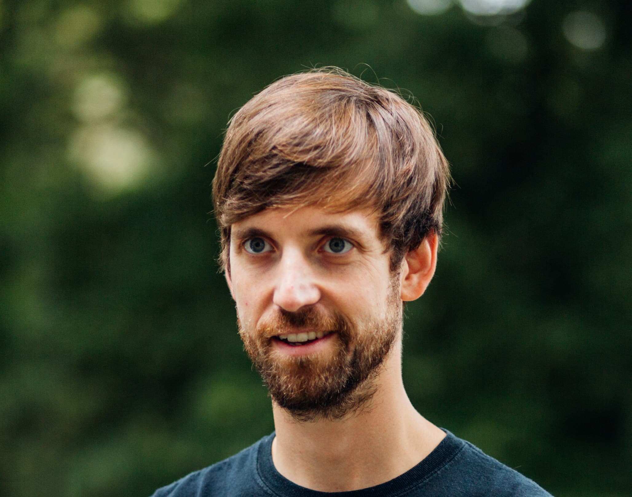 A man with short brown hair and a beard is looking slightly to the side and smiling. He is wearing a black T-shirt. The background is a blurred outdoor setting with green foliage.