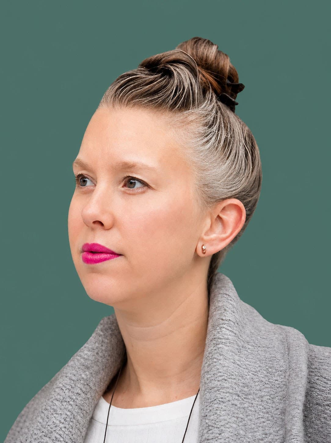 A person with light skin and gray hair in a top bun, wearing bright pink lipstick, a gray coat, and a white top, looks slightly to the left against a solid teal background.
