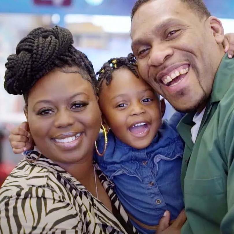 A joyful family of three smiles at the camera. The mother, with braided hair, wears a patterned top. The father, in a green shirt, embraces their daughter who has beaded braids and is dressed in blue. All three look very happy.