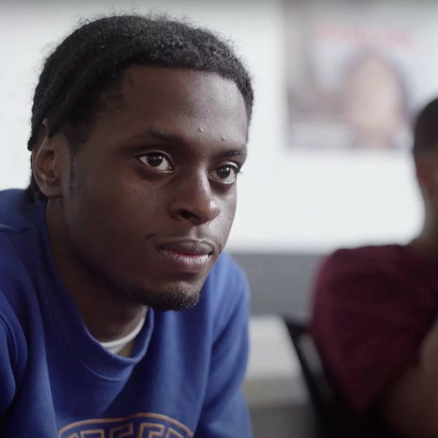 A young man with short braids and a mustache is attentively listening during a discussion. He is wearing a blue sweatshirt. In the blurred background, another person is partially visible, sitting in a maroon shirt. The setting appears to be an indoor room.