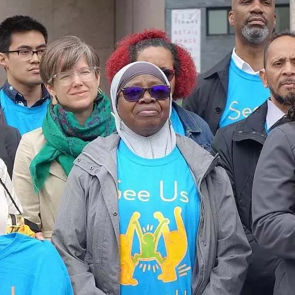 A group of people stands together, most wearing blue shirts with yellow graphics and the words "See Us." They appear to be participating in an outdoor event or rally. The central person wears a gray jacket, white headscarf, and sunglasses. People are smiling and attentive.