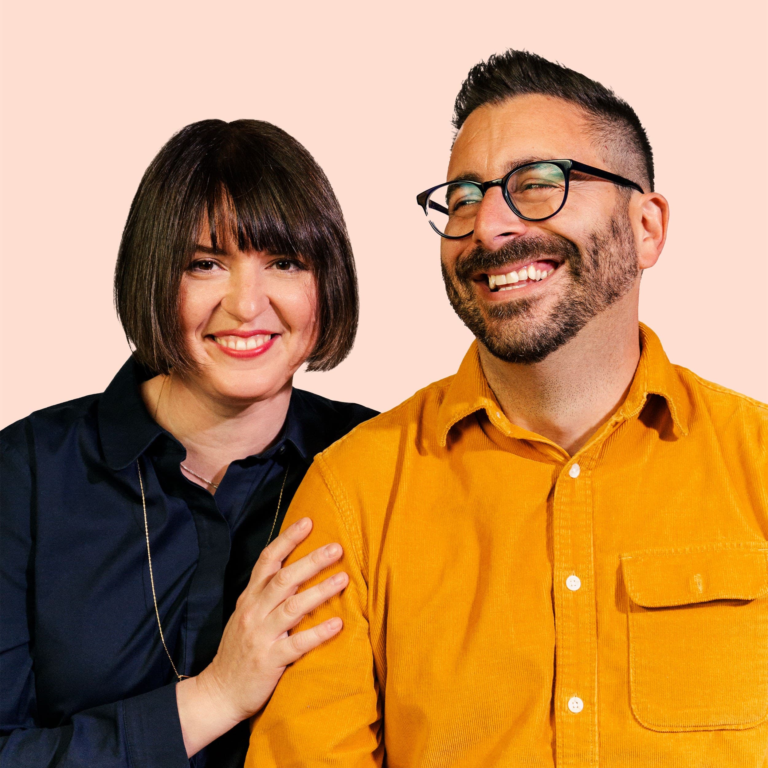 A woman with a bob haircut and a man with a beard and glasses smile together. She wears a dark shirt and rests her hand on his shoulder. He wears a mustard-yellow shirt. The background is a light peach color.