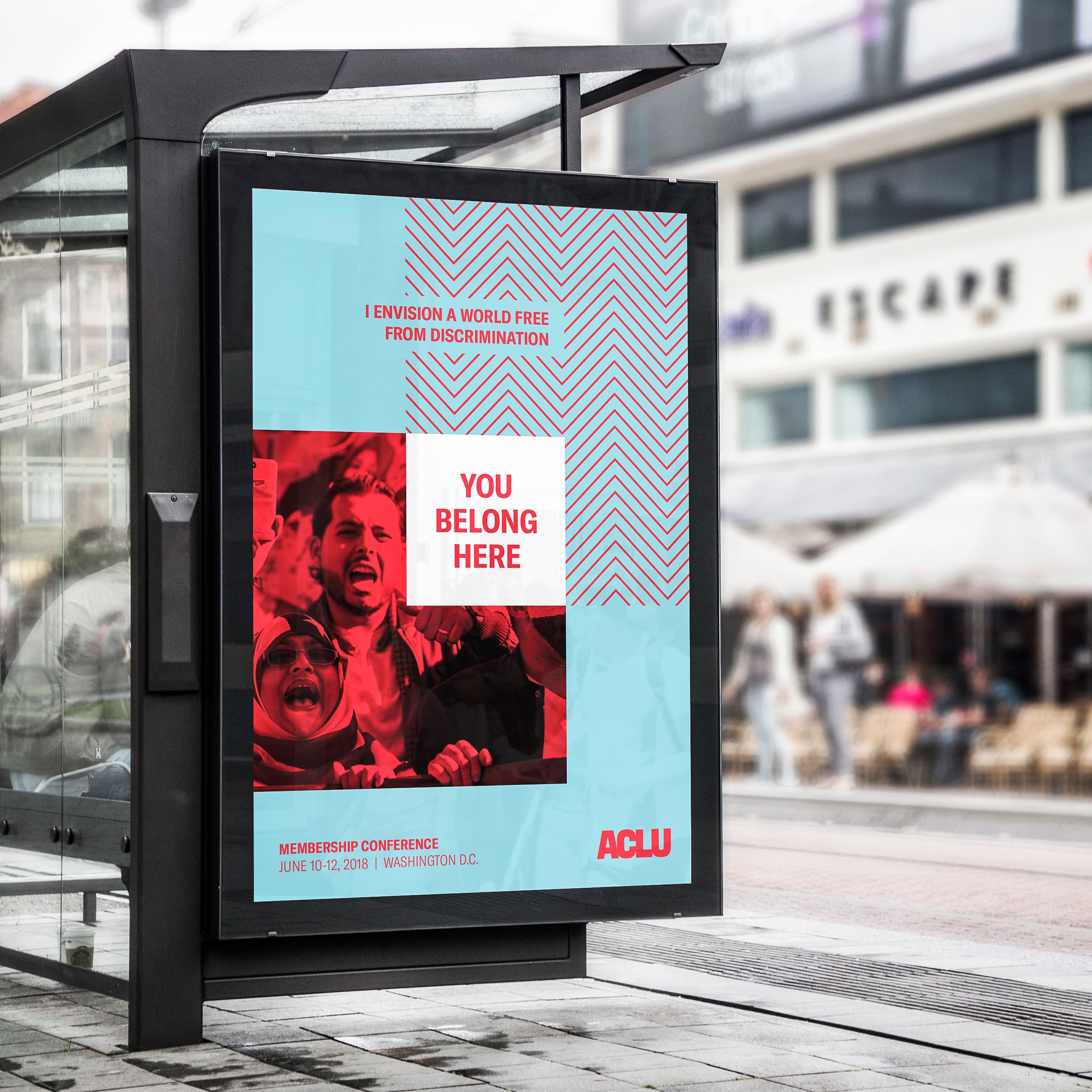 A bus stop billboard displays an ACLU advertisement featuring a red and white geometric design with a photo of smiling people. The text reads, "I envision a world free from discrimination. You belong here." It promotes the membership conference on June 10-12, 2018, in Washington, D.C.