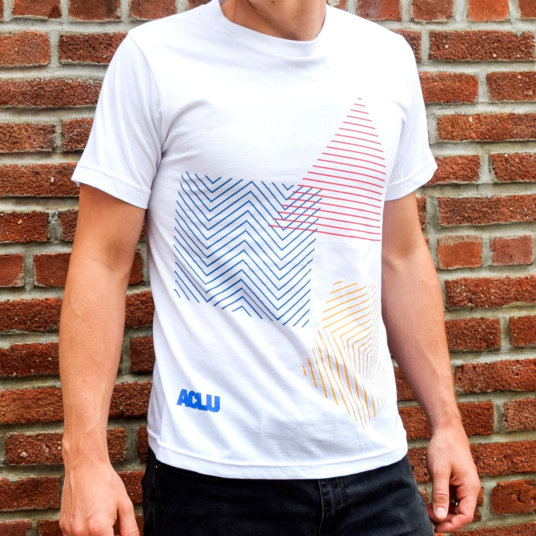 A person is wearing a white t-shirt with geometric shapes in blue, red, and yellow. The letters "ACLU" are printed in blue on the lower left side of the shirt. The person is standing against a brick wall. Their face is not visible in the image.