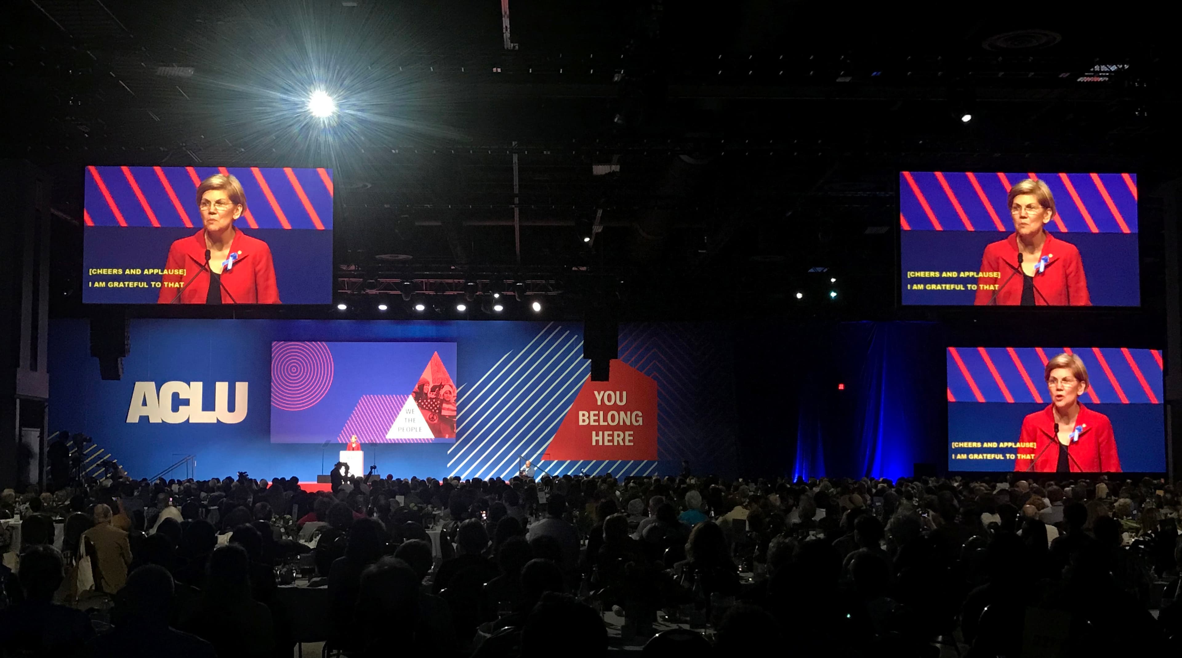 A large auditorium is filled with people attending an ACLU event. On stage, a speaker dressed in red is addressing the audience. Large screens above display a close-up of the speaker. The backdrop features "ACLU" on the left and "YOU BELONG HERE" on the right.