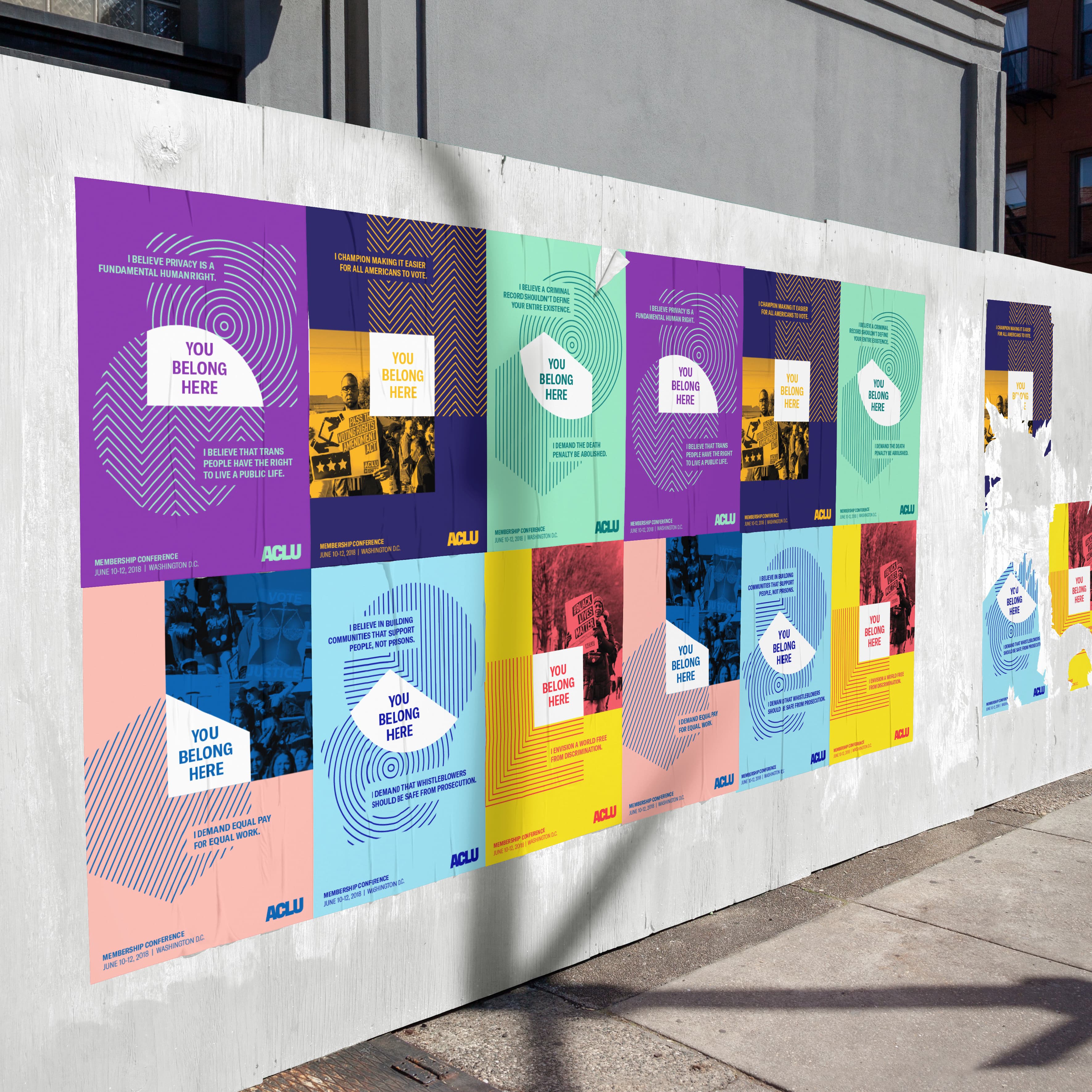 A street wall is covered with colorful posters displaying the message "YOU BELONG HERE" in bold text. Each poster features fingerprints as a background design, with varying colors such as purple, orange, green, pink, and blue. The ACLU logo is visible on all posters.