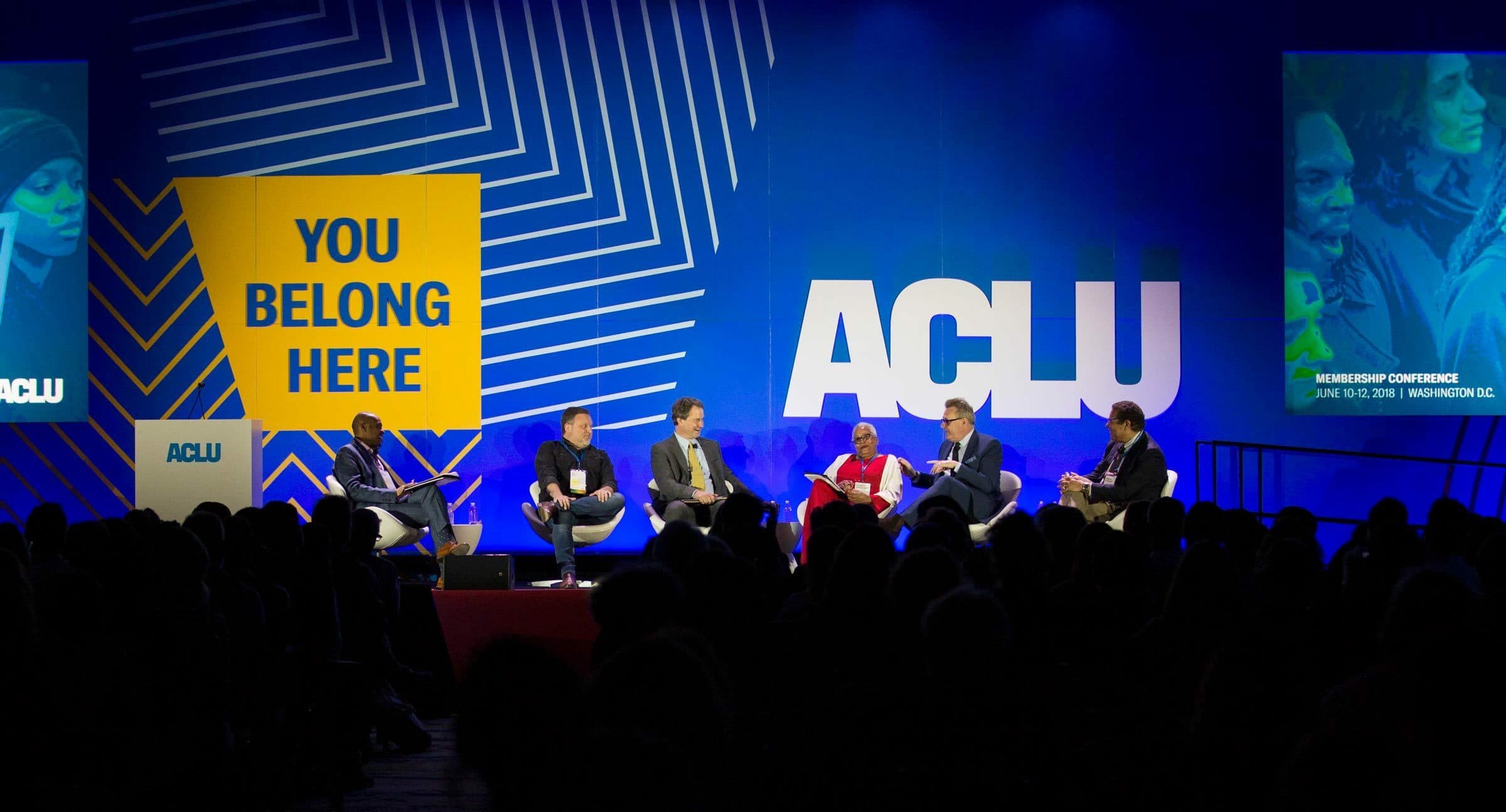 A group of people sit on a stage in a panel discussion at a conference. The large screen behind them displays "ACLU" and a sign reads "YOU BELONG HERE." The backdrop is blue with white diagonal lines and additional text about the event is visible.