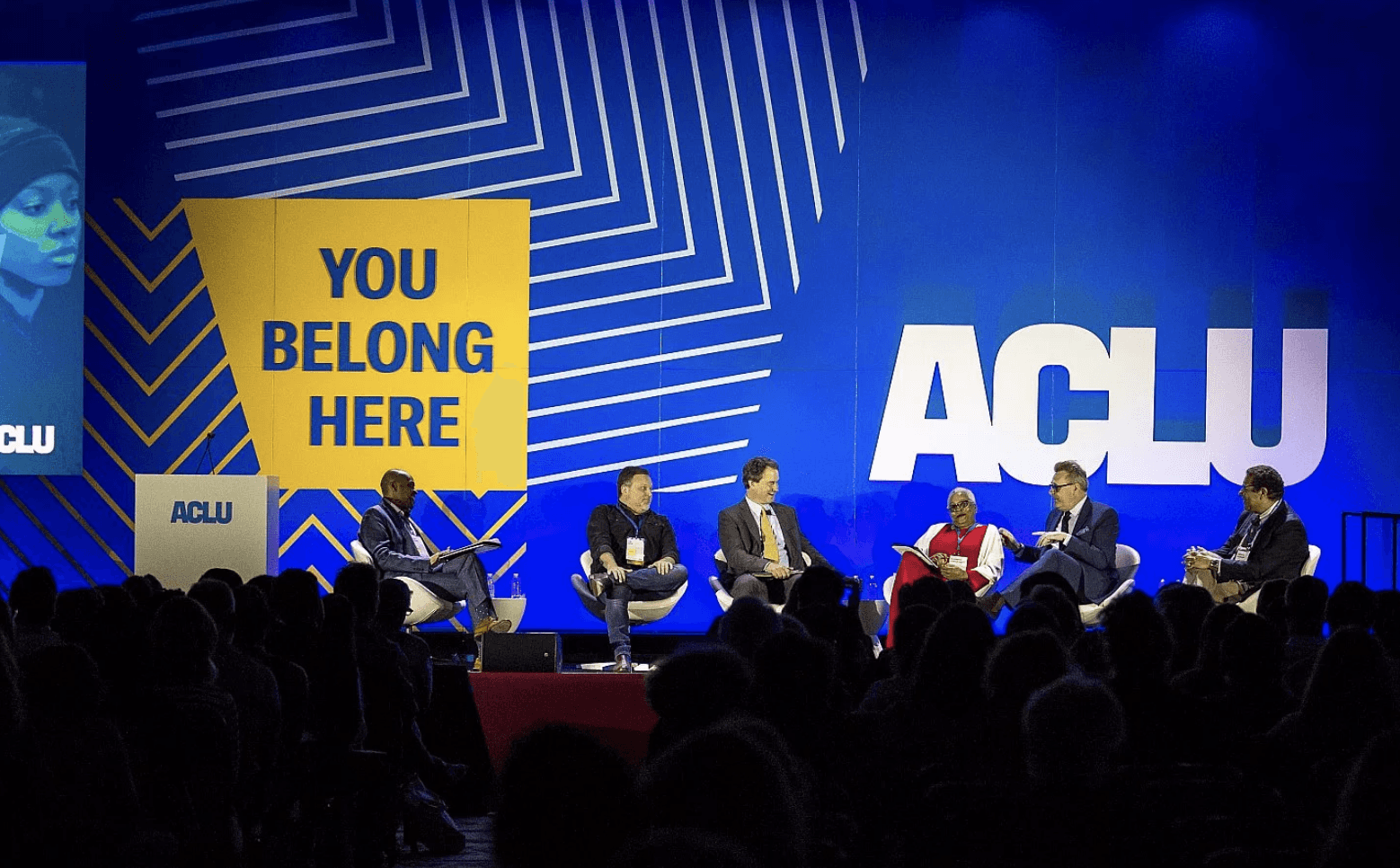 A panel of six people engage in a discussion on a stage with the ACLU logo prominently displayed in the background. A large yellow sign reads "YOU BELONG HERE." An audience is partially visible in the foreground. The backdrop features a blue and white geometric design.