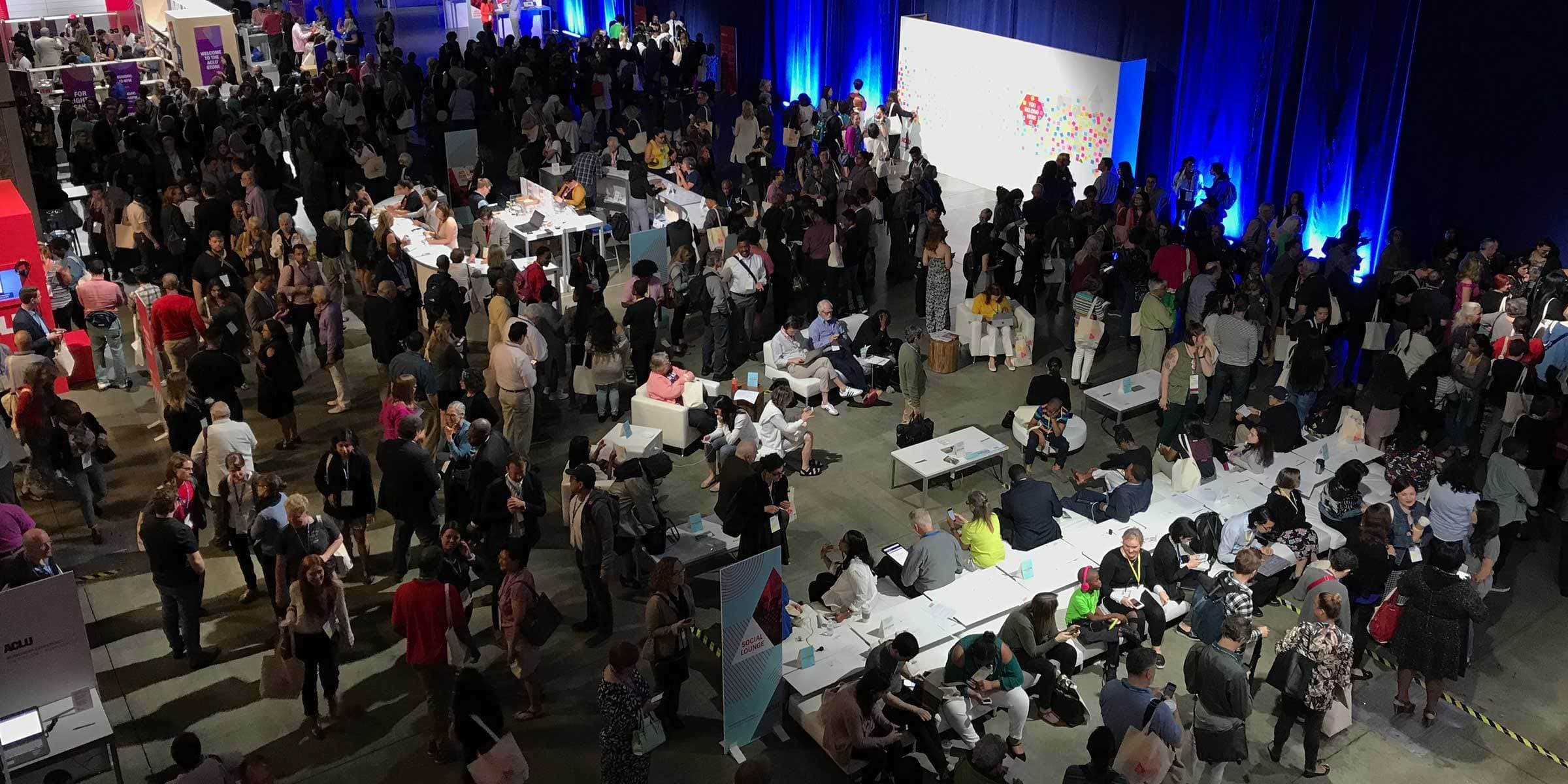 A crowded convention hall filled with people attending various booths and sitting at tables. Many individuals are standing, conversing, and engaging with the exhibitors, while others are seated in small groups. The setting is lively and brightly lit with colorful displays.