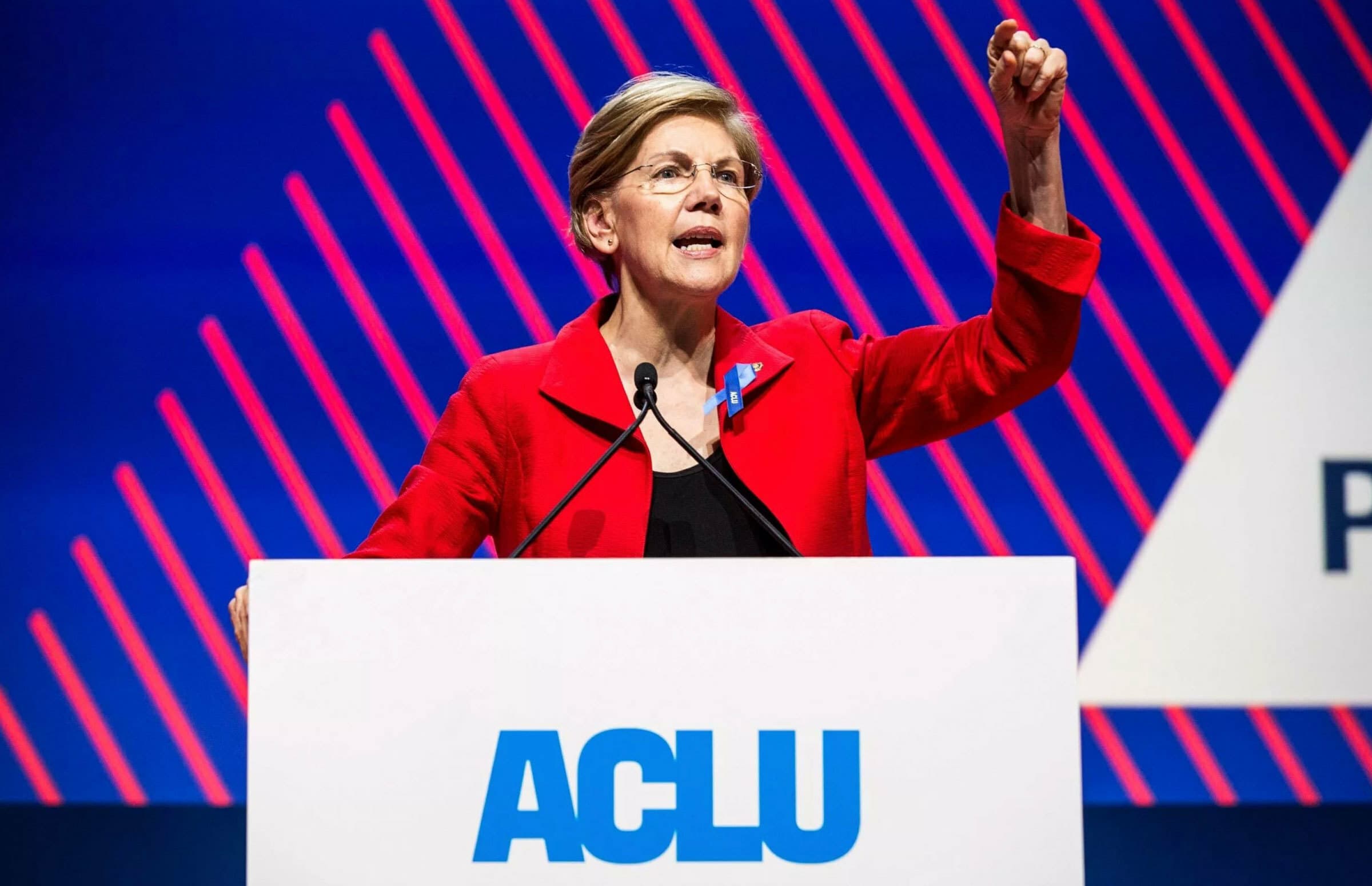 A person in a red blazer speaks passionately at a podium with the letters "ACLU" in blue on it. The background features diagonal red and blue stripes with partial text visible. The individual is raising one hand, appearing to emphasize a point.