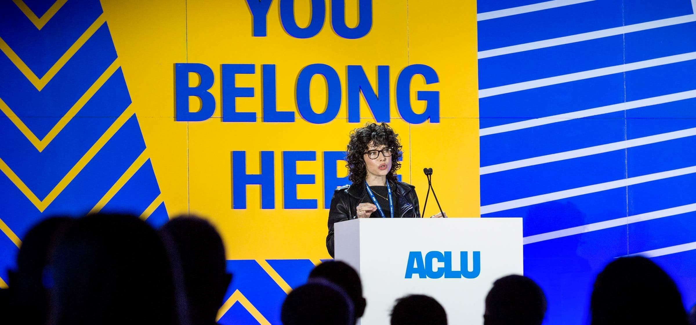 A person stands behind a podium with the ACLU logo, addressing an audience. The backdrop features the text "YOU BELONG HERE" in large, bold letters, accompanied by geometric patterns in yellow and blue. Silhouettes of audience members are visible in the foreground.