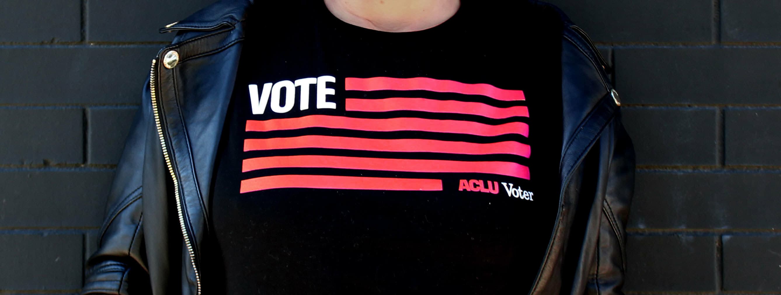 A person wearing a black leather jacket and a black shirt with the word "VOTE" in white, and the ACLU Voter logo featuring red stripes resembling the American flag. The background consists of dark, horizontal bricks.