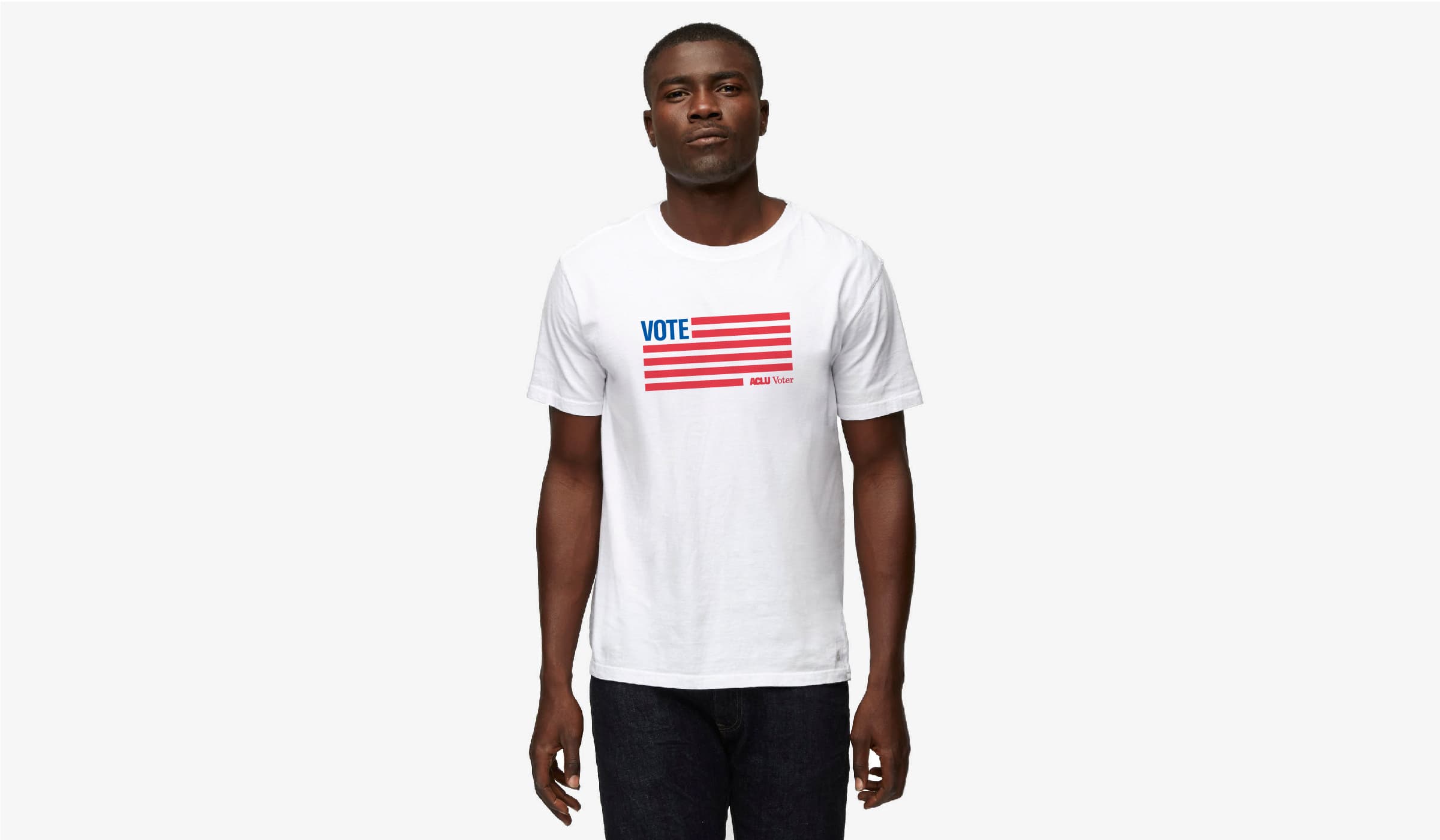 A man is wearing a white T-shirt featuring a design of an American flag with "VOTE" written in blue above the red stripes. The man stands against a plain white background, facing forward with a neutral expression.