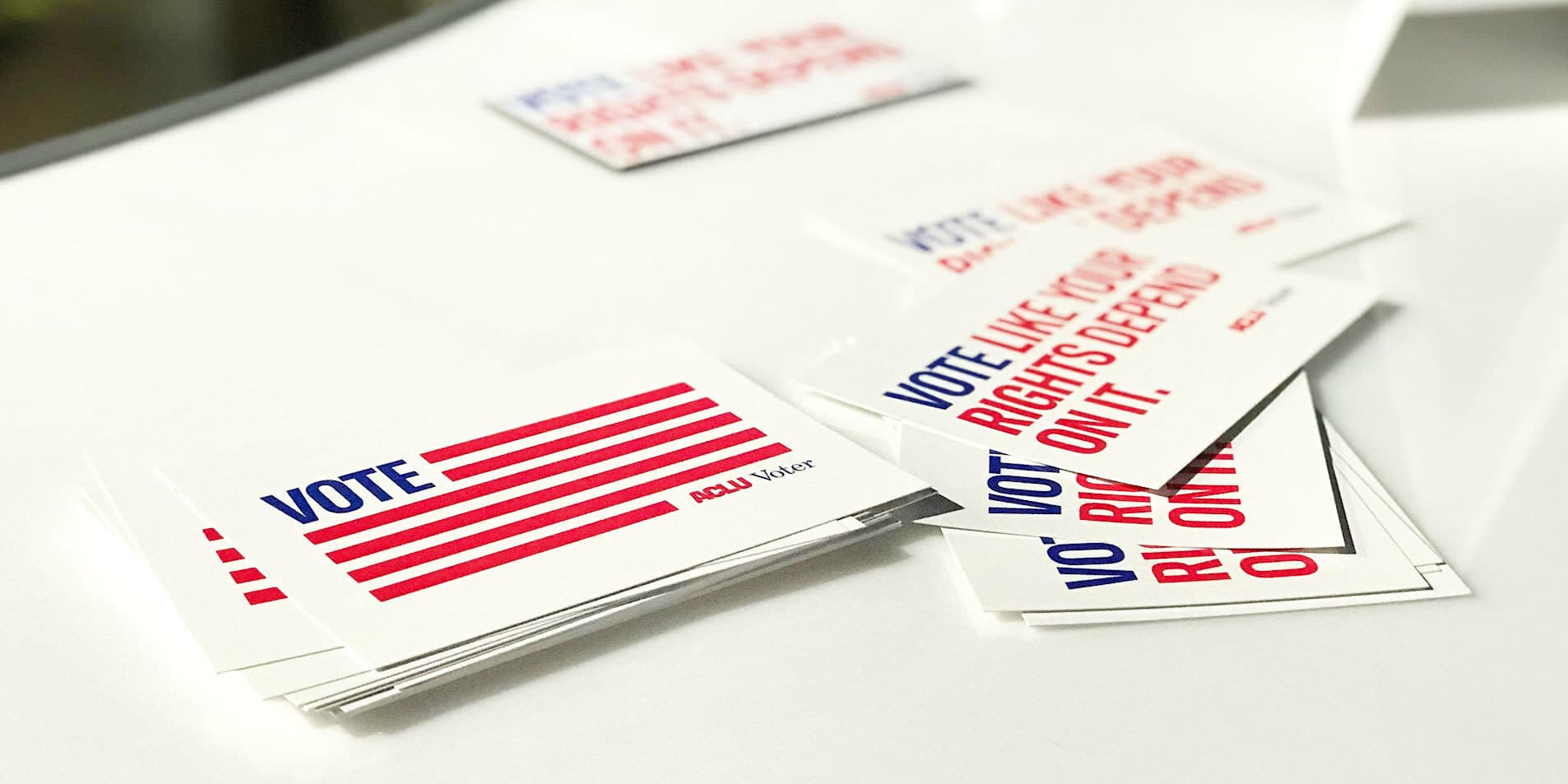 Stacked cards with the word "VOTE" in bold blue letters and red stripes resembling an American flag, along with the ACLU logo, are displayed on a white surface. Mixed among them are other cards with the phrase "VOTE LIKE YOUR RIGHTS DEPEND ON IT.