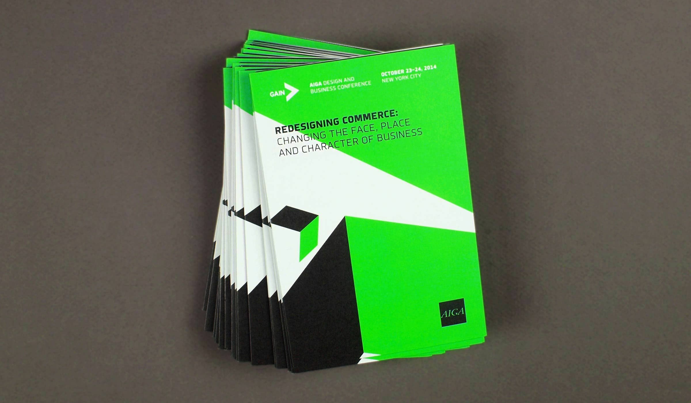 A stack of green and black booklets lies against a gray background. The top booklet reads "Redesigning Commerce: Changing the Place, Place, and Character of Business" with event details at the top: "AIGA GAIN Design and Business Conference, October 23-24, 2014, New York City.