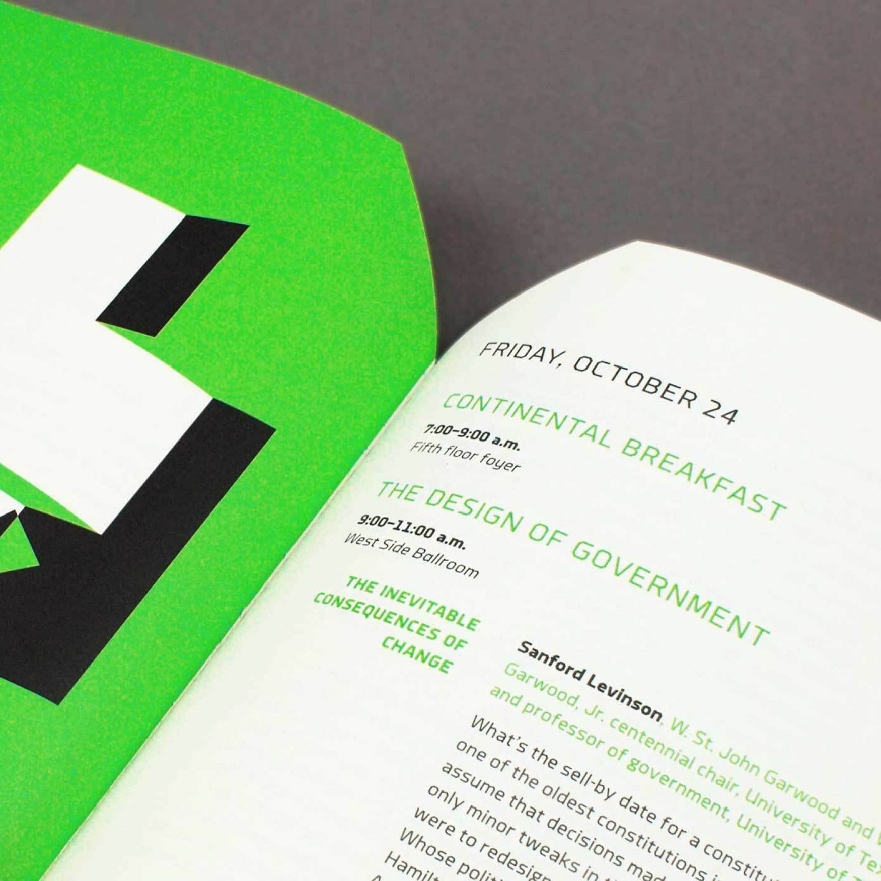 Close-up of an open book on a gray surface, showing a page titled "Continental Breakfast" and "The Design of Government" on Friday, October 24. The schedule lists times, locations, and a speaker named Sanford Levinson. The page has a green and white color scheme.