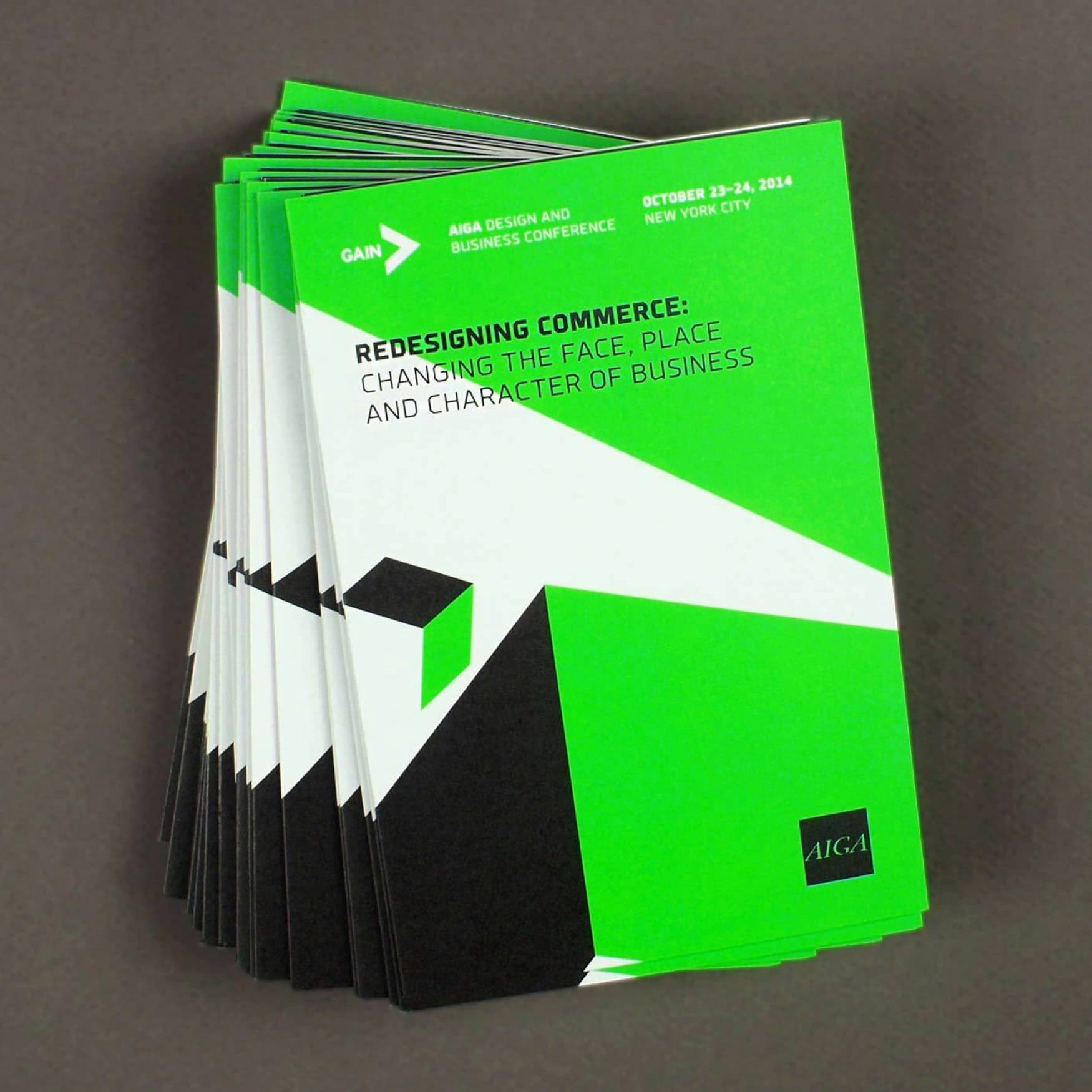A stack of green and black conference booklets titled "Redesigning Commerce: Changing the Face, Place, and Character of Business." Text includes "AIGA Design and Business Conference" and "October 23-24, 2014, New York City." The AIGA logo is visible.