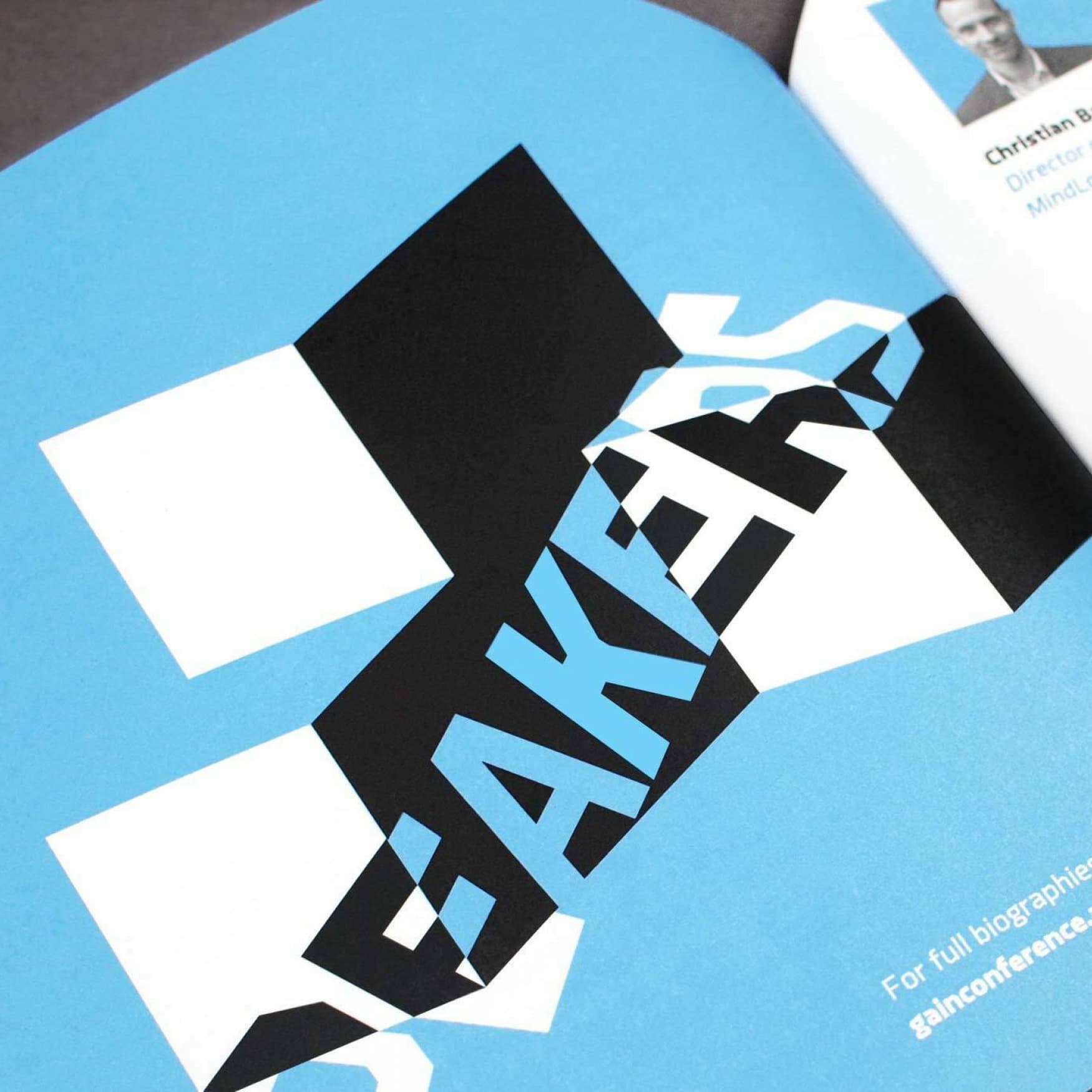 Close-up of a blue and white booklet page with the word "SPEAKERS" prominently displayed in large, black, geometric text. A section of a headshot and brief information about one of the speakers is visible at the top right corner of the image.