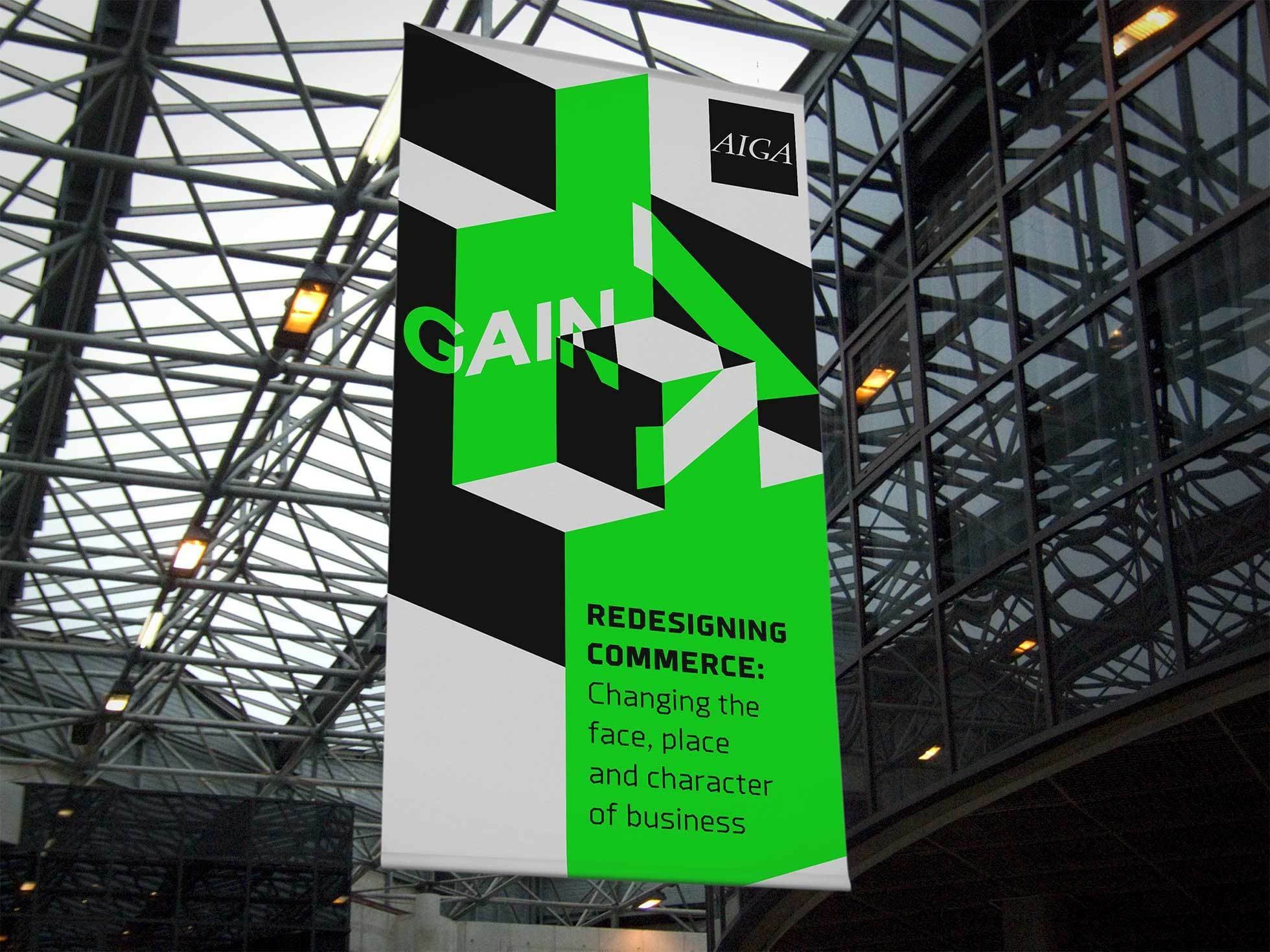 A vertical banner hanging in a modern atrium with glass ceilings and large windows. The banner features geometric designs and the text "GAIN" and "REDESIGNING COMMERCE: Changing the face, place, and character of business" on a green and black background. The AIGA logo is at the top.