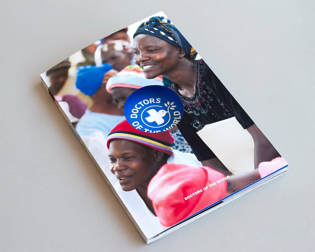 A brochure titled "Doctors of the World" lies on a grey surface. The cover features smiling individuals wearing colorful clothing and headscarves. The prominently visible logo consists of a white cross inside a blue circle with the organization's name.
