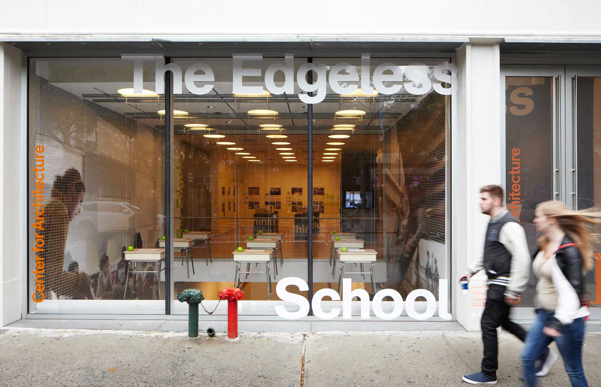 A street view of The Edgeless School, housed in a glass-front building with visible classrooms inside. The signage on the window reads "The Edgeless School" and includes the name "Center for Architecture." Two people walk past the building.