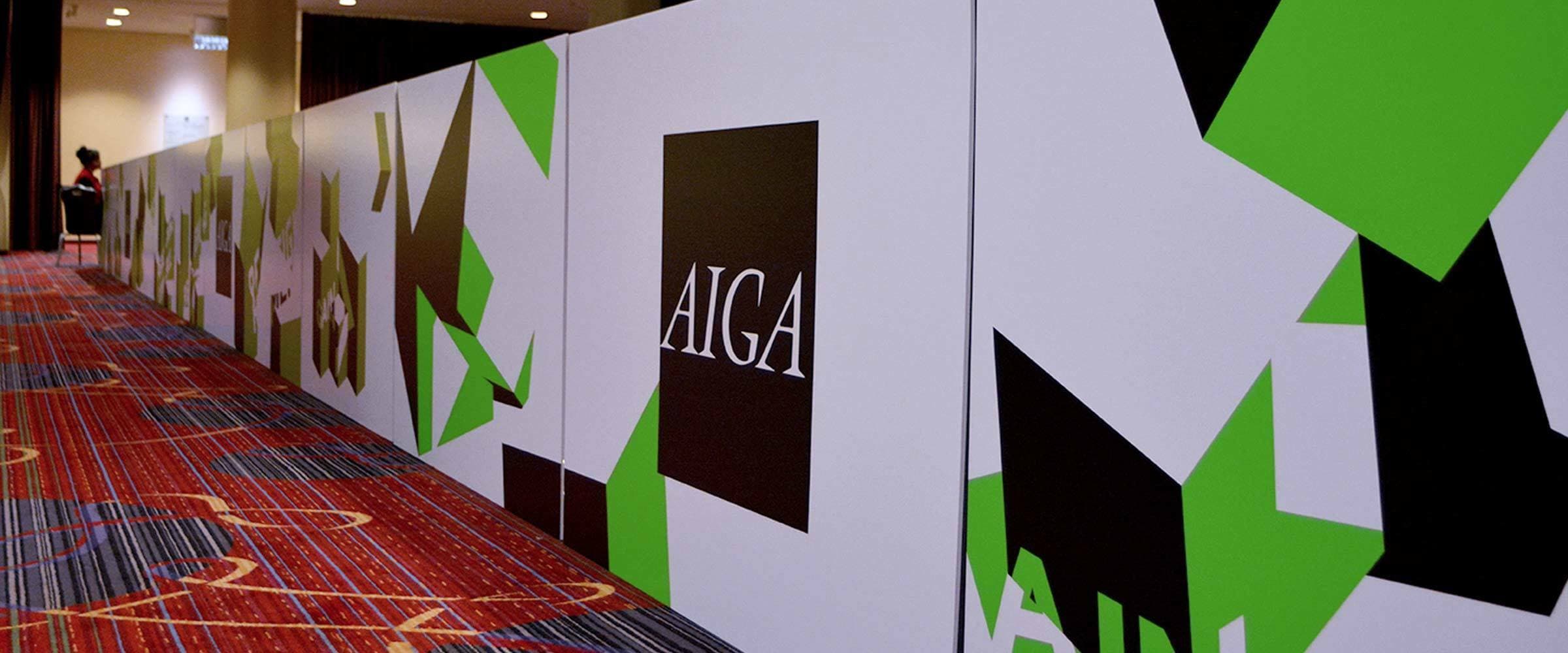 A long wall features a geometric design with black and green shapes on a white background. The letters "AIGA" are prominently displayed in a black square in the center. The wall is in a carpeted area with a person visible in the background.