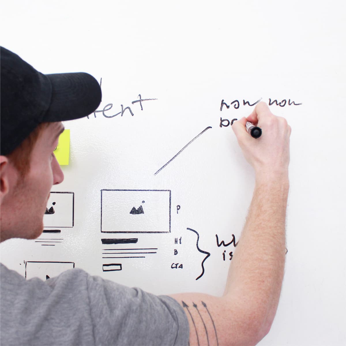 A person in a black cap and gray T-shirt writes on a whiteboard with a black marker. The board features various sketches, including website wireframes, notes, and scribbles. The person has a tattoo on their right arm.