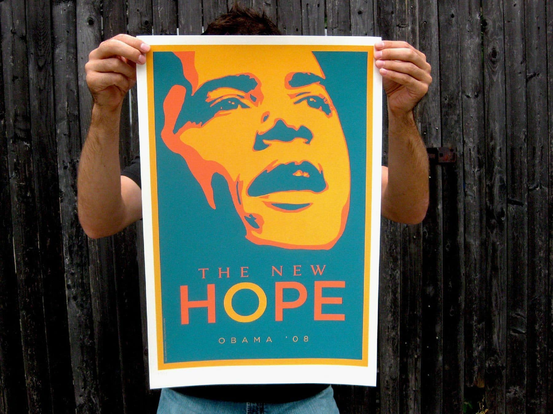 A person holds up a poster of Barack Obama in a bold, colorful style with the text "The New Hope" and "Obama '08". The background of the image is a weathered wooden wall.