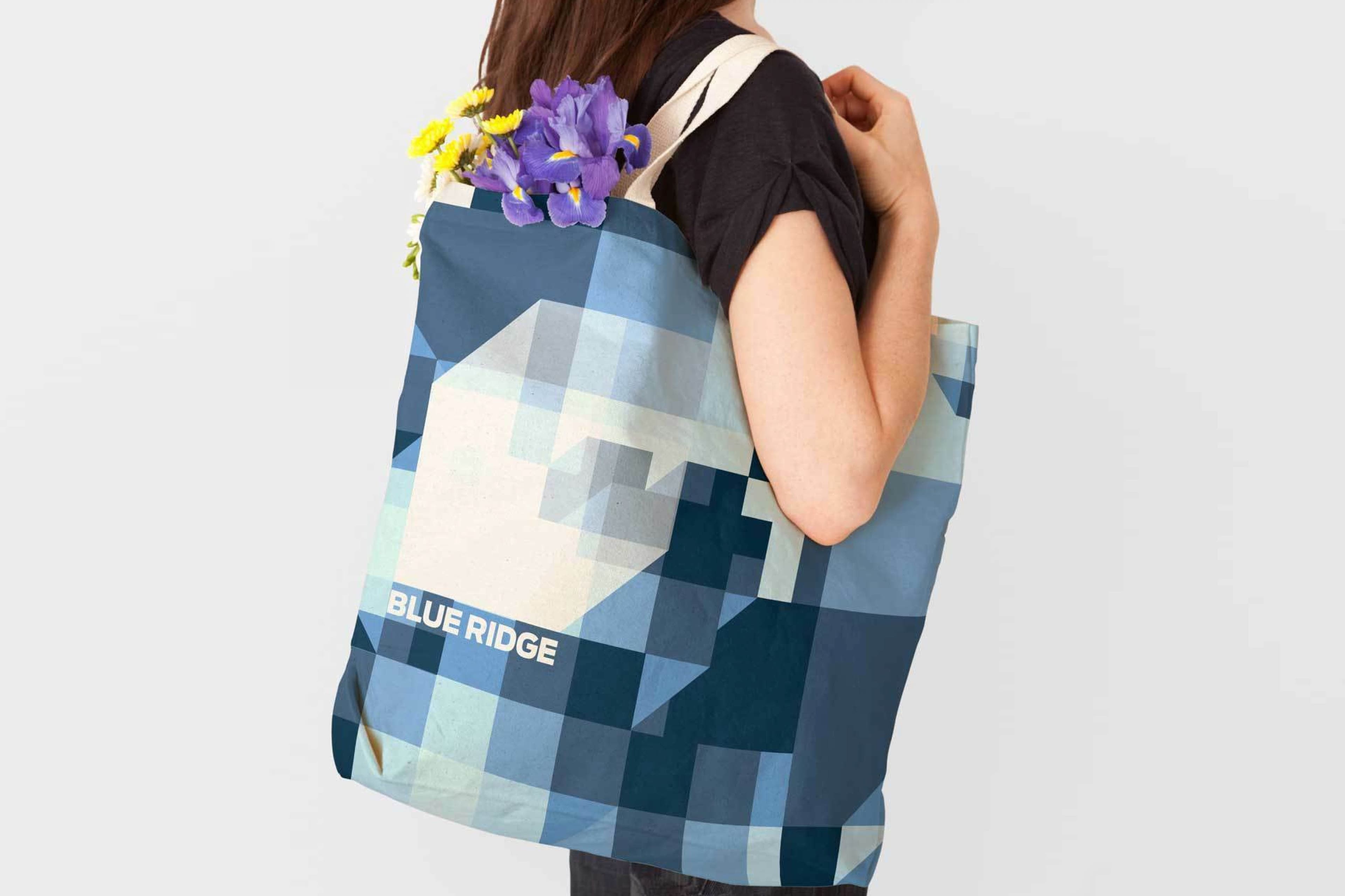 A person carrying a tote bag with a geometric blue and gray pattern and the text "BLUE RIDGE." The bag is over their shoulder and filled with vibrant yellow and purple flowers. The person is wearing a black shirt and their face is not visible.