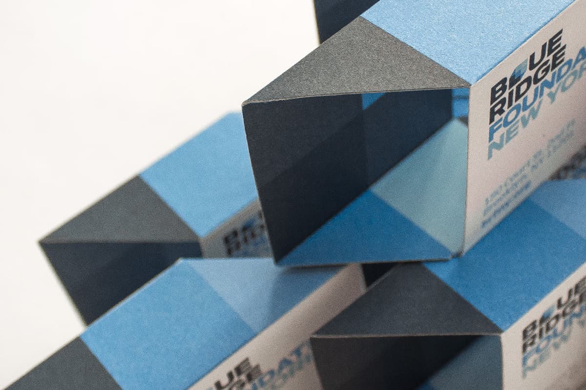 A close-up photograph shows a geometric arrangement of blue, black, and white folded paper shapes. The paper shapes appear to be rectangular with hollow centers, creating a layered and interlocking pattern. Text reading "BLUE RIDGE PRINTING NEW YORK" is partially visible.