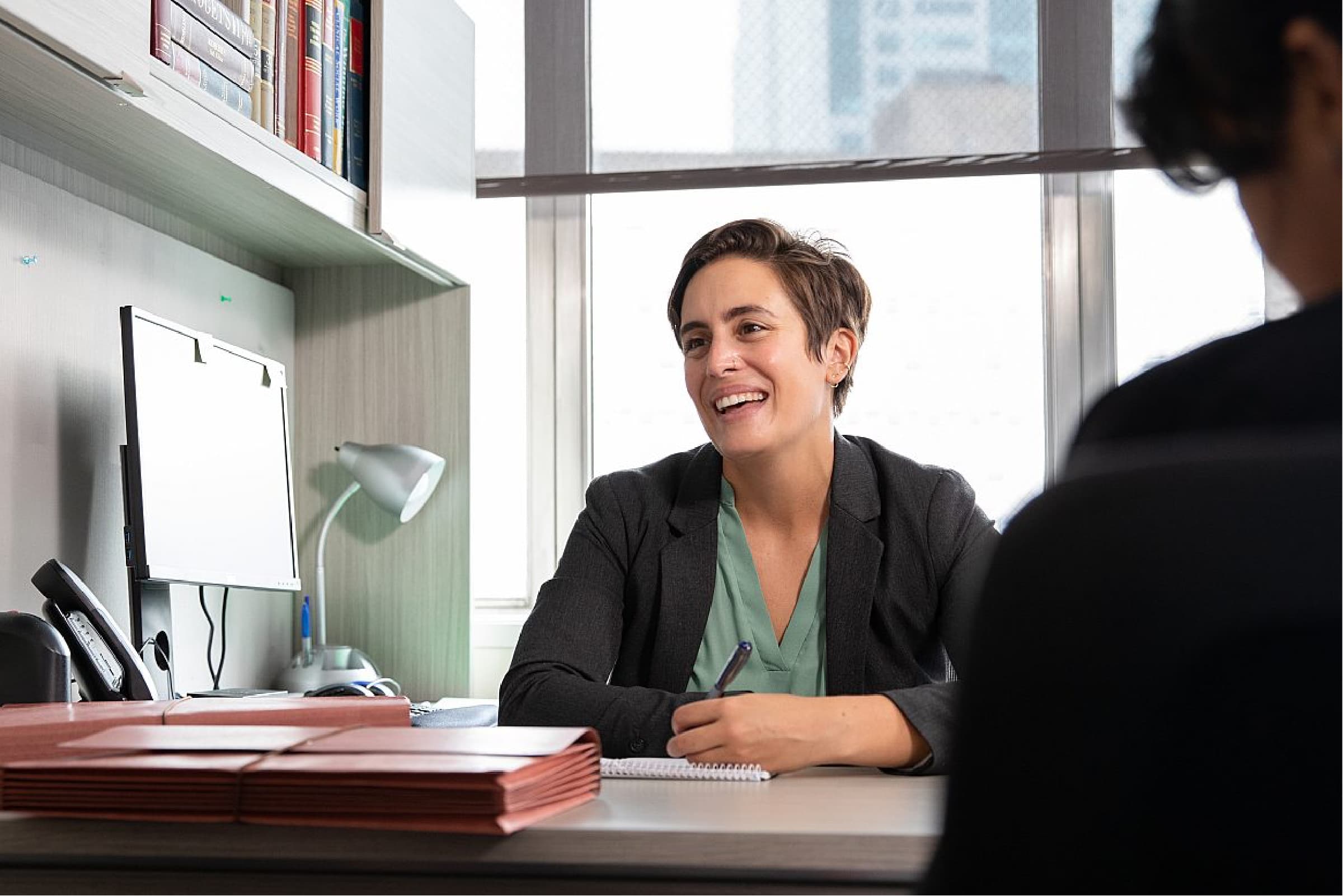 A person with short hair, wearing a blazer, is sitting at a desk in an office, smiling and holding a pen. The desk has files, a computer monitor, a phone, and various other office supplies. Another person, blurred, is sitting across from them.