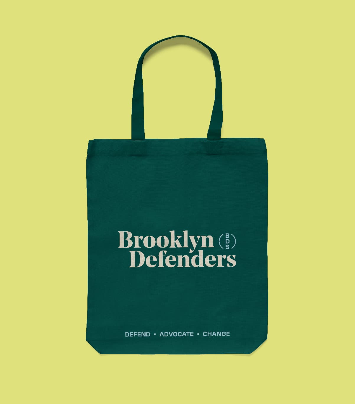 A dark green tote bag on a light green background, displaying the text "Brooklyn Defenders" in large, white font. Below, smaller text reads "Defend • Advocate • Change." There is also a vertical circular logo to the right of "Defenders.