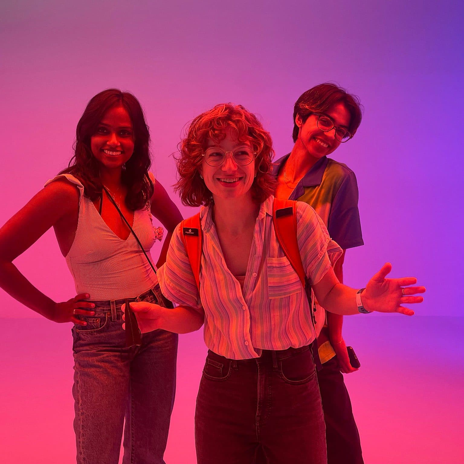 Three people standing in a brightly lit space with colorful lighting. They are smiling and appear to be enjoying themselves. One person at the center is wearing glasses and a striped shirt, the person on the left is in a tank top, and the person on the right in a short-sleeve shirt.