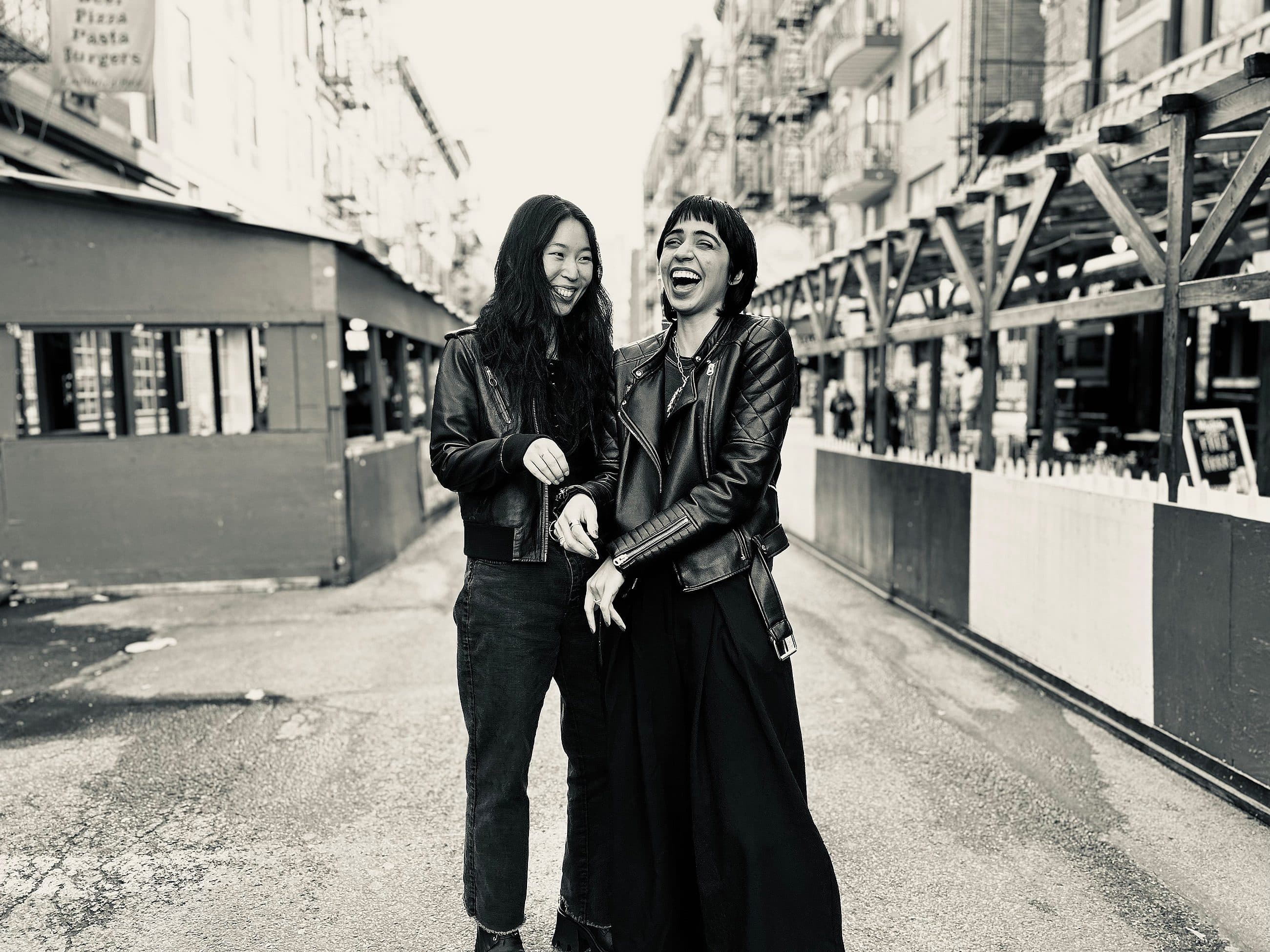 Two women stand laughing together on an urban street. Both are wearing dark clothing with leather jackets. The street is lined with buildings and outdoor structures. The scene is captured in black and white, highlighting the joyful moment shared between them.