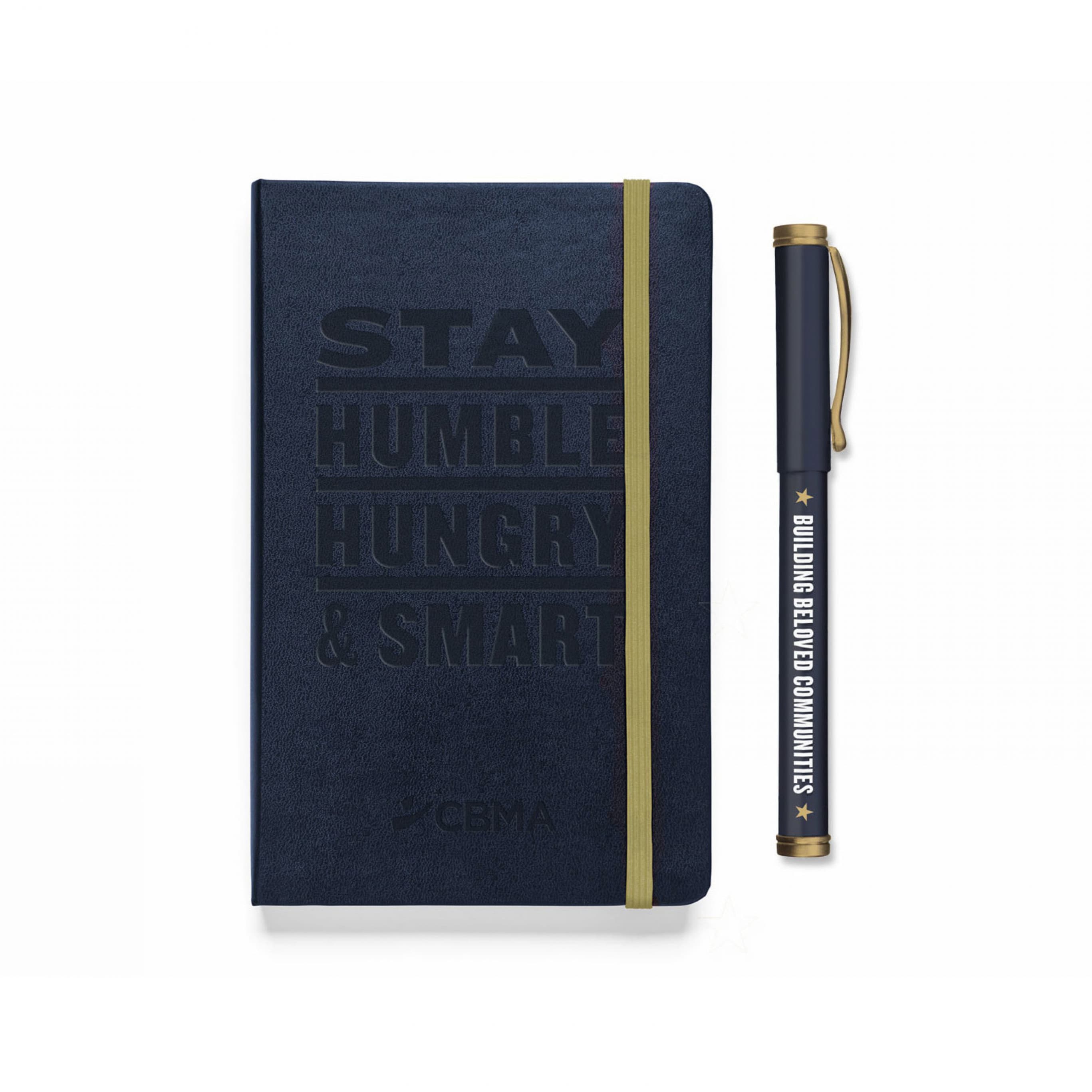 A navy blue notebook with the text "STAY HUMBLE HUNGRY & SMART" debossed on the cover, along with the acronym "CBMA" and a small logo at the bottom. The notebook has a yellow elastic band. Next to it is a black pen with the text "BUILDING BELOVED COMMUNITIES.