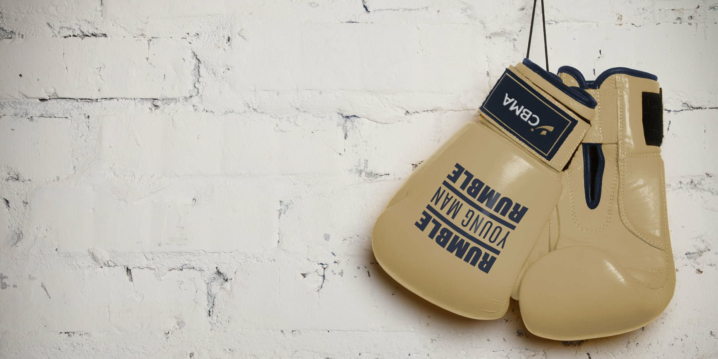 A pair of beige boxing gloves hang from a black rope against a white brick wall. The gloves are branded with the inscriptions "Rumble Young Man Rumble" and "Combat" along with a logo.