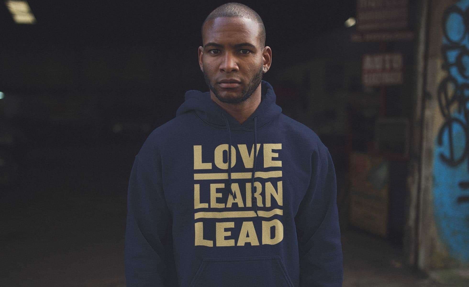A person with short hair and a beard is wearing a navy blue hoodie with the words "LOVE LEARN LEAD" printed in bold yellow capital letters. The background is dimly lit with some graffiti visible on the right side and a building in the shadows.