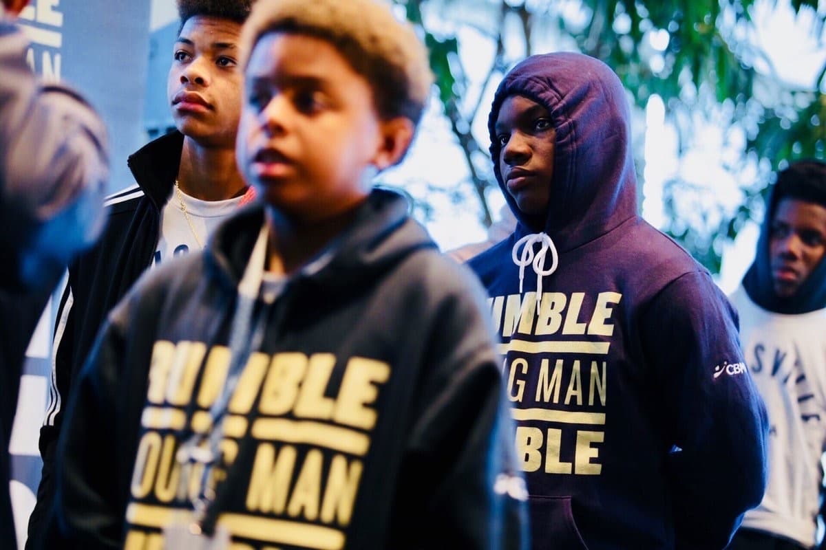 A group of young men, some wearing hoodies that read "HUMBLE YOUNG MAN HUMBLE," stand attentively. One wears a beige headband, and another has a hood covering most of his head. They are indoors, with blurred greenery visible in the background.