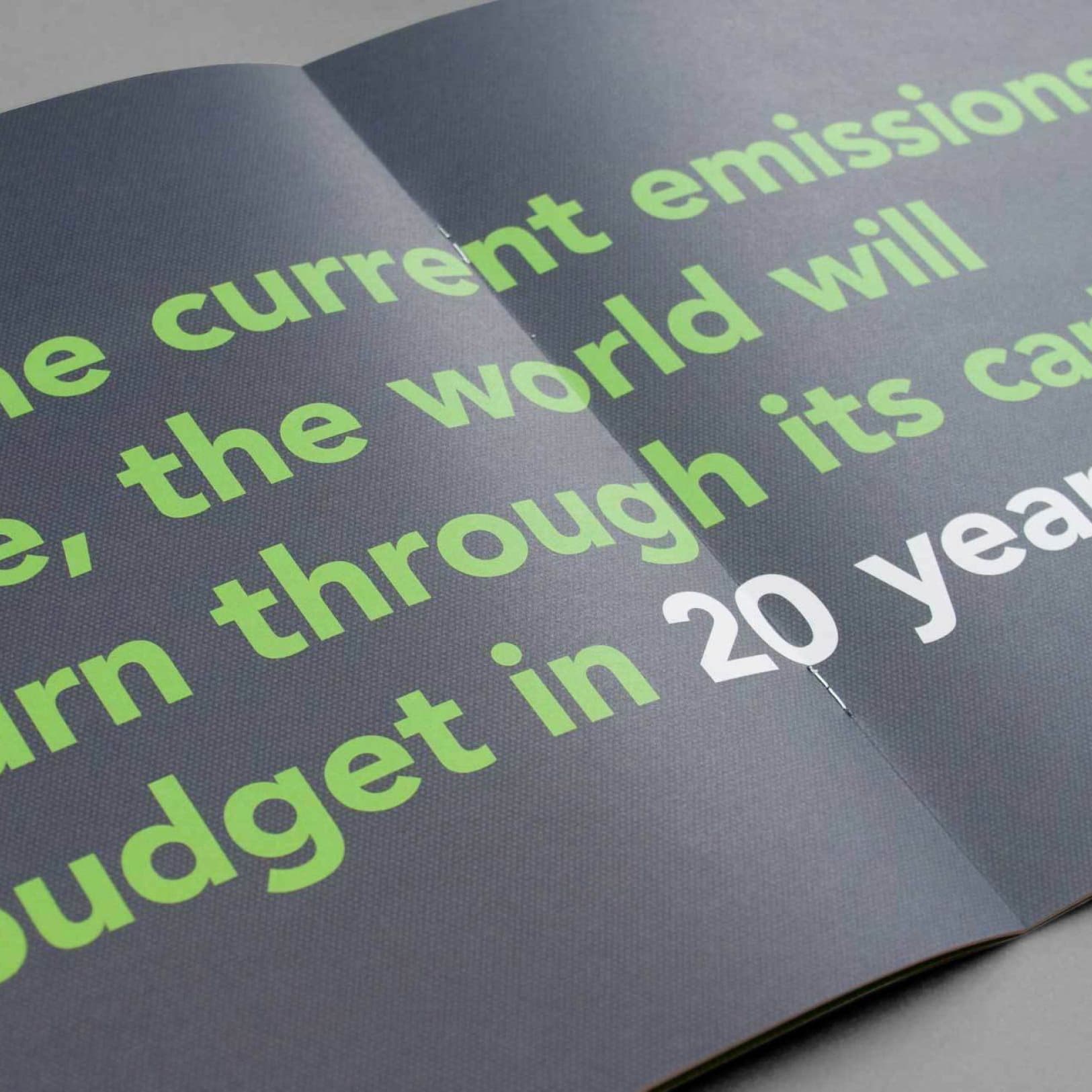 Close-up of a page in a booklet with text in large, bold green and white letters. The text reads: "the current emissions rate, the world will burn through its carbon budget in 20 years." The background is dark grey, and the text is spread across two pages.