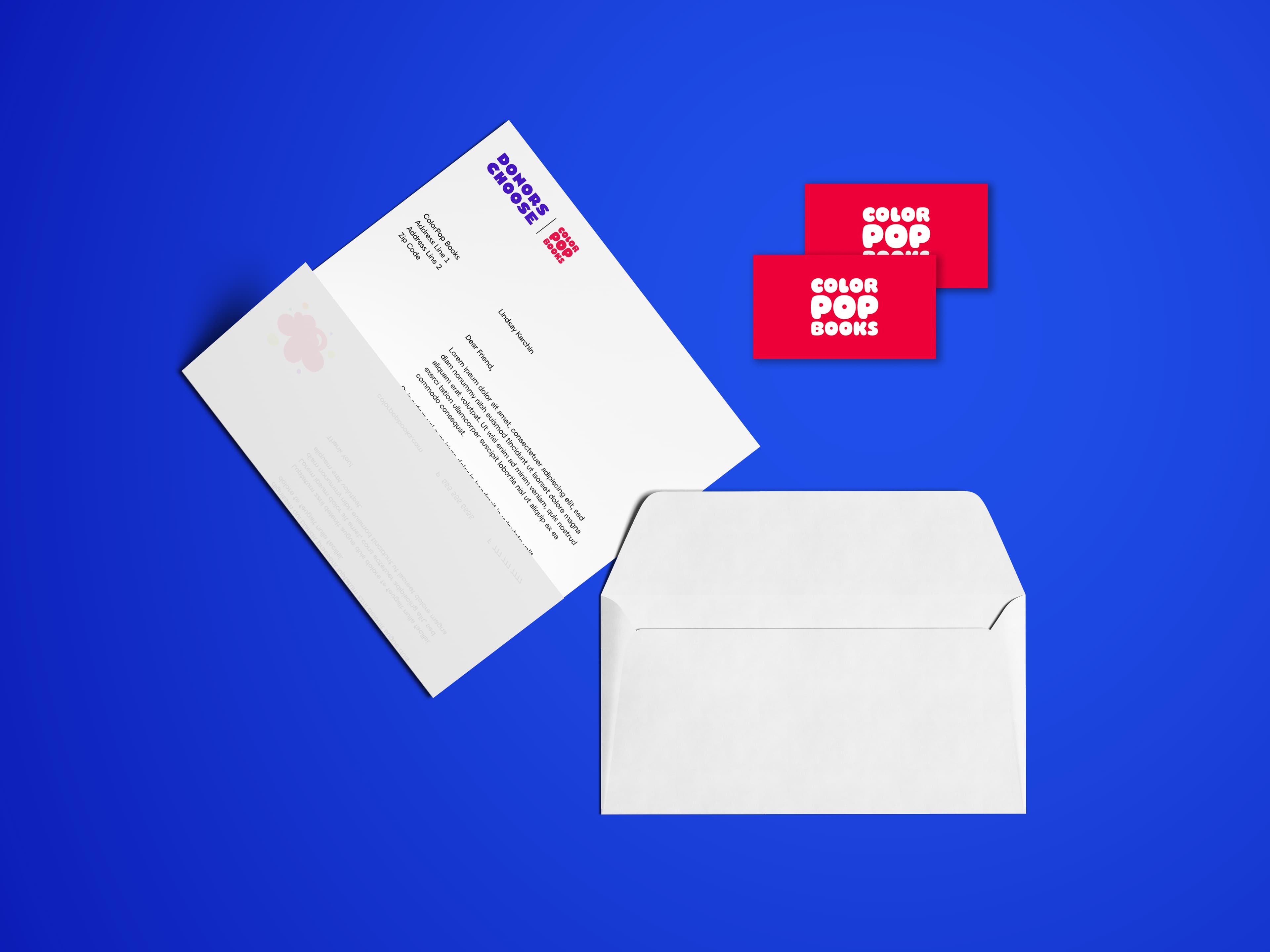 A flat lay on a blue surface showing two red business cards with "Color Pop Books" in white text, a white letterhead with the same logo, and an address, alongside an open white envelope.