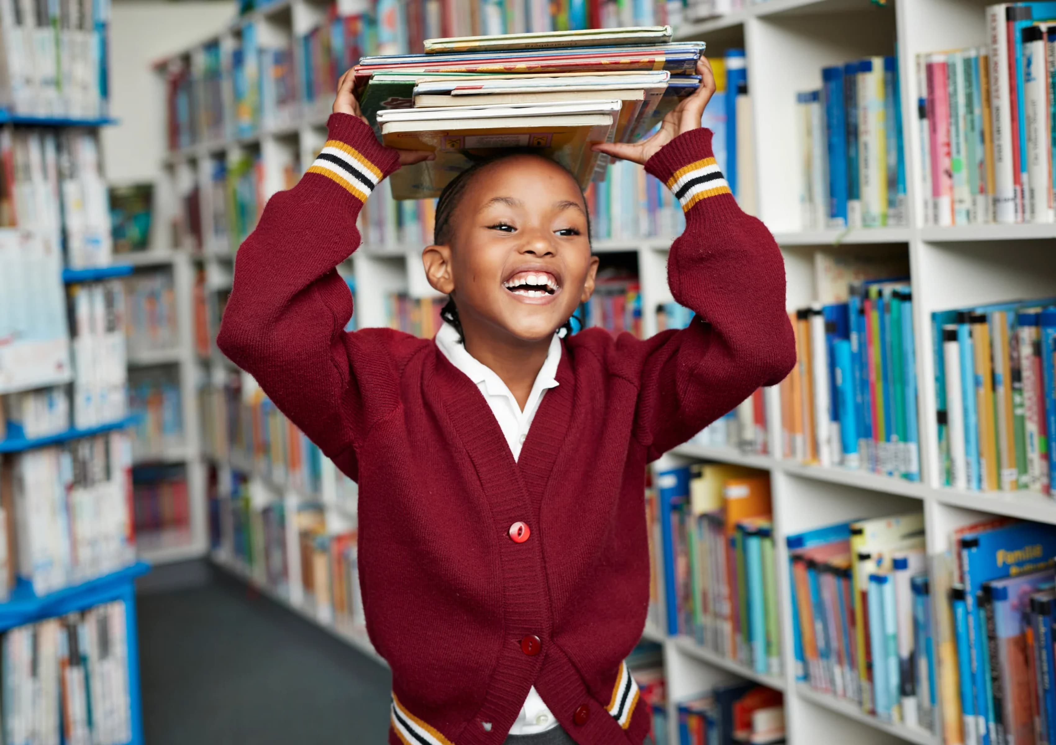 A child stands in a library, smiling brightly while balancing a stack of books on their head. They are wearing a maroon cardigan with striped cuffs, and the shelves behind them are filled with colorful books.