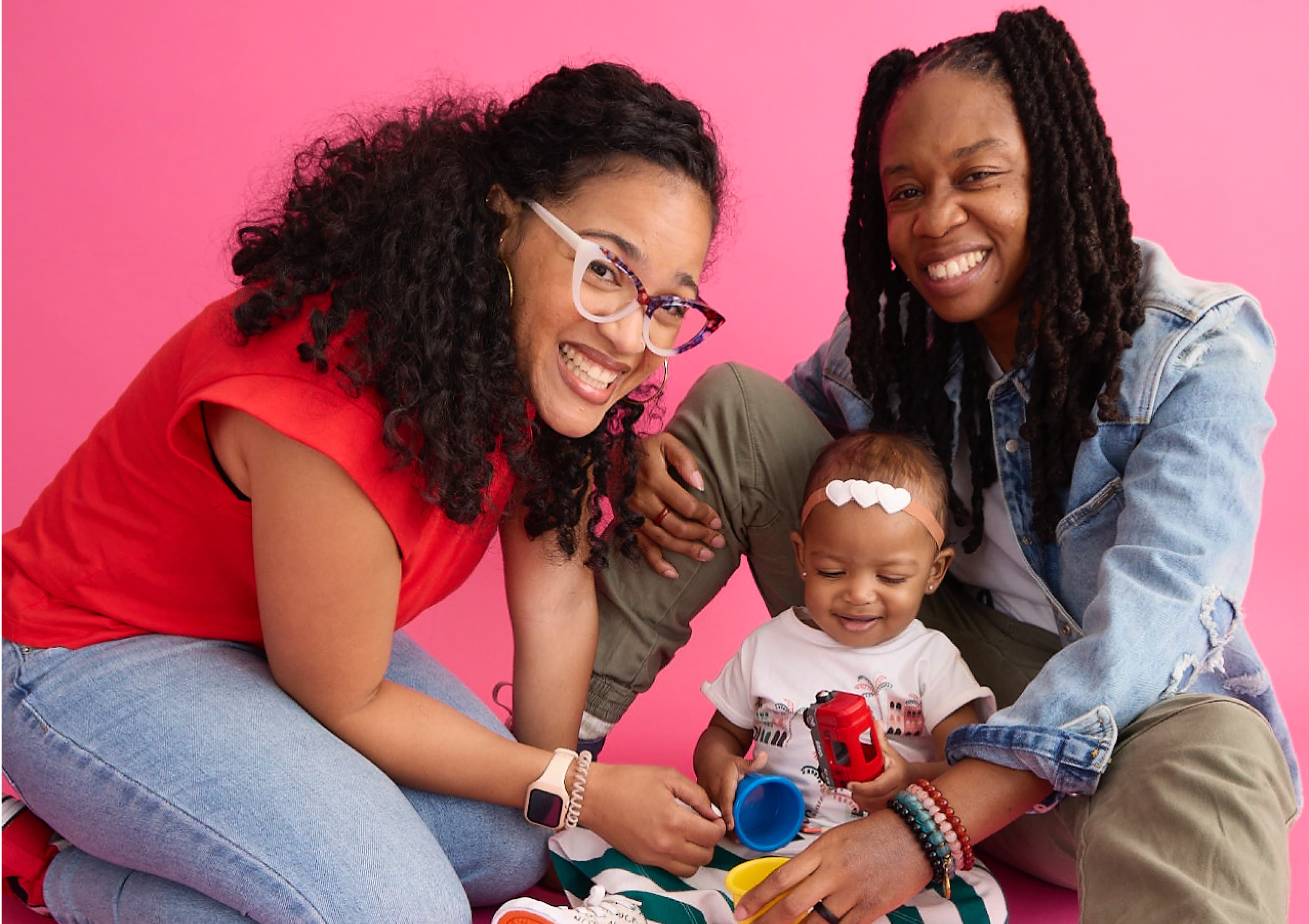 A happy family of three sits together against a pink background. Two adults, one in glasses and a red shirt, the other in a denim jacket, smile warmly at the camera. A cheerful baby, wearing a headband and holding toys, sits between them on a striped blanket.