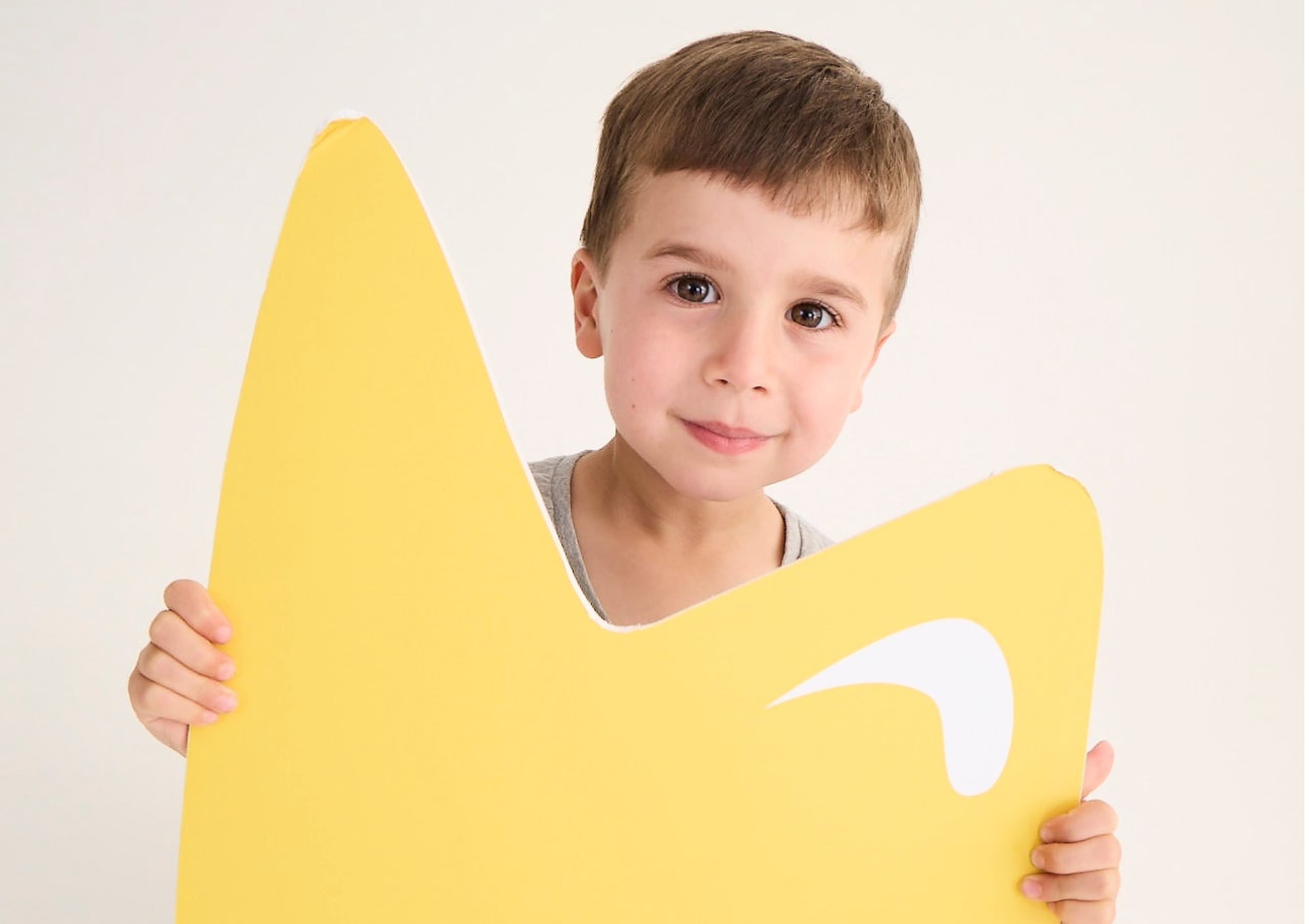 A young child with short brown hair holds a large, yellow cardboard cutout in the shape of a crown against a plain white background. The child smiles softly, and the crown has a white decorative element on it.