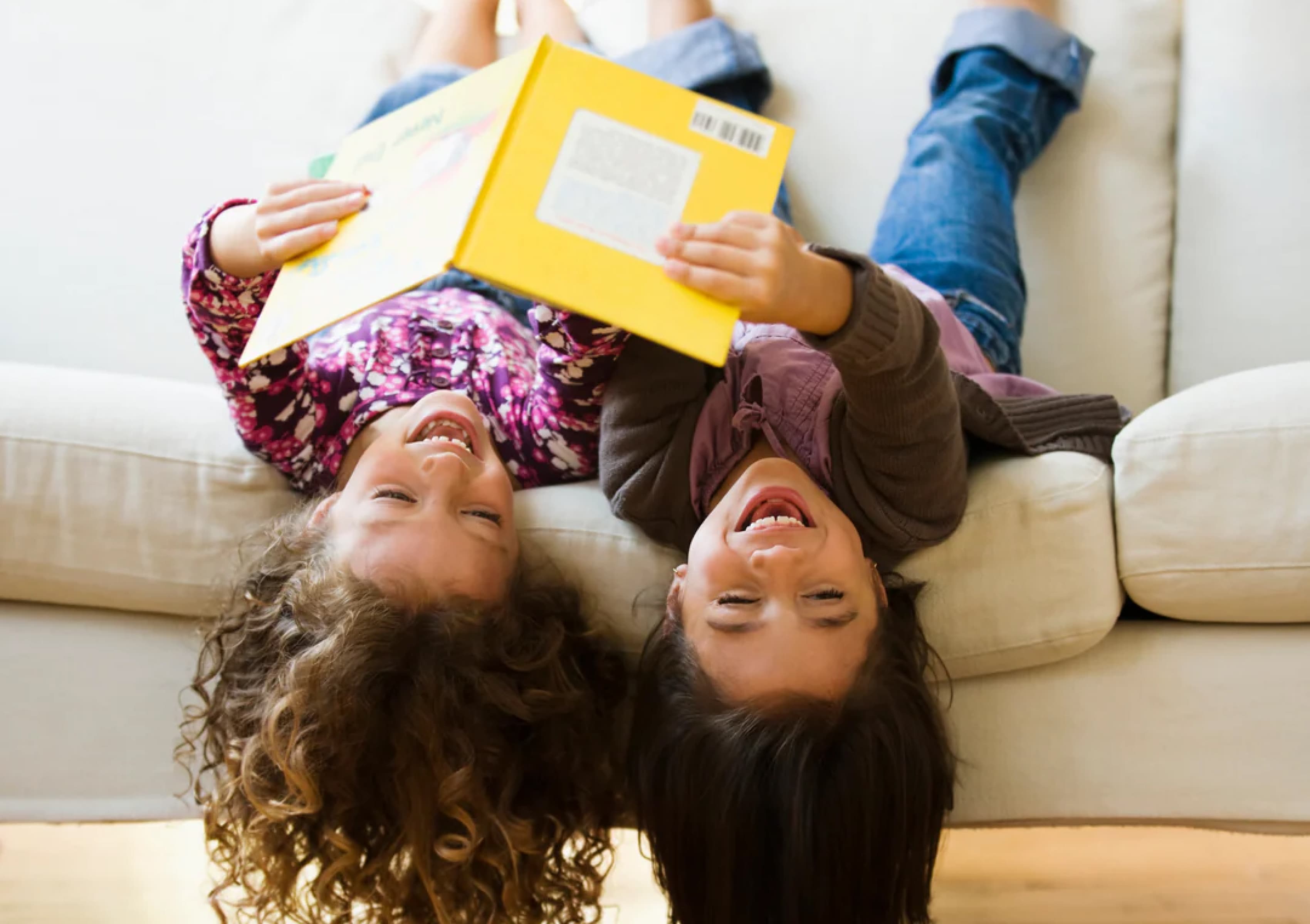 Two children are lying upside down on a couch, their heads hanging over the edge, and laughing joyfully while holding and reading a yellow book together. One child has curly hair and wears a floral shirt, while the other has straight hair and wears a brown sweater.