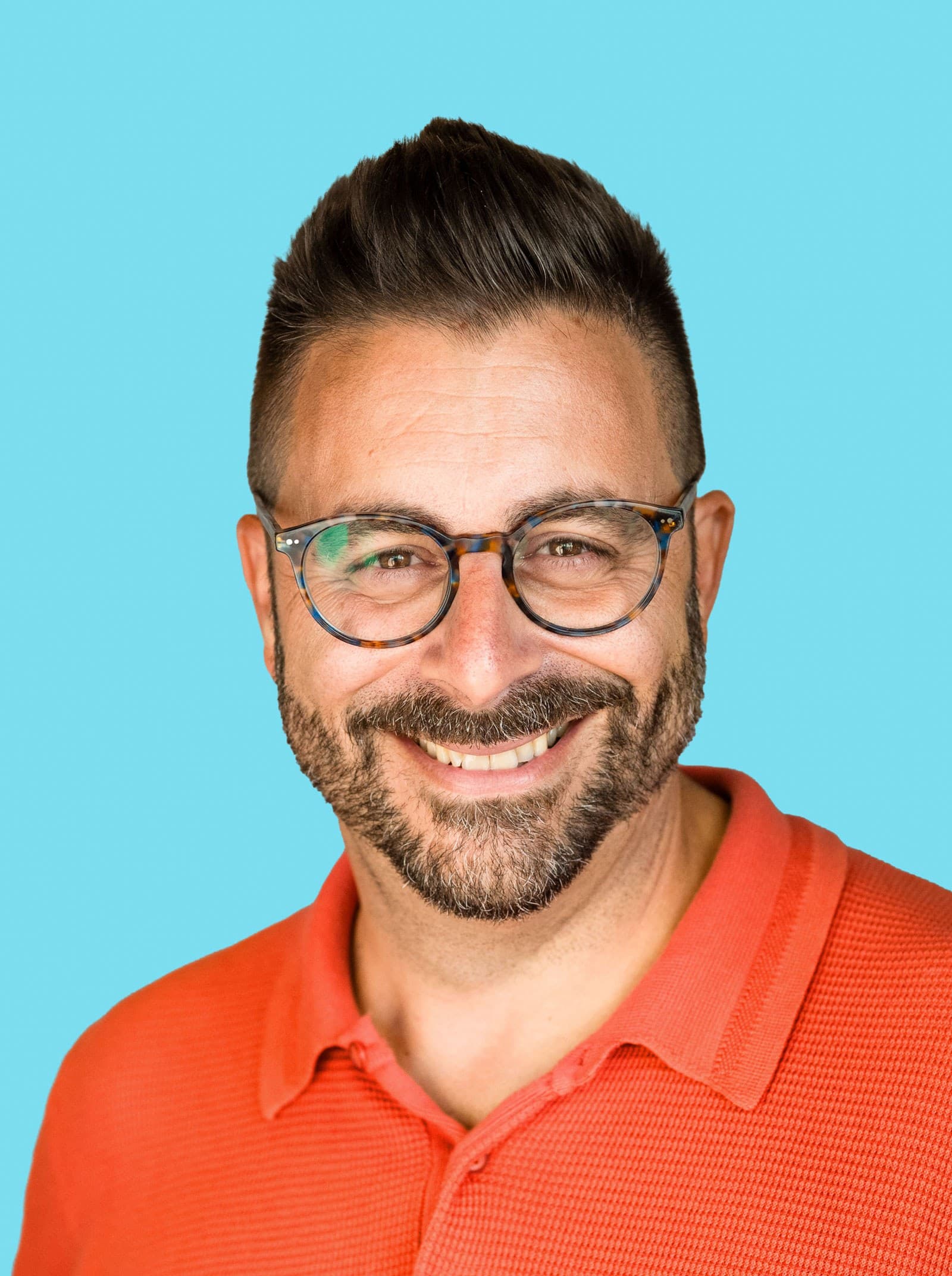 A smiling man with short dark hair, a beard, and glasses wears a bright orange polo shirt against a solid light blue background.