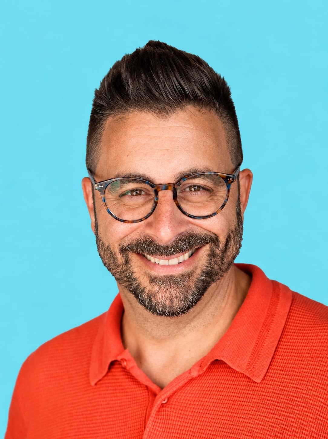 Smiling man with short dark hair, beard, and glasses wearing an orange collared shirt against a solid light blue background.