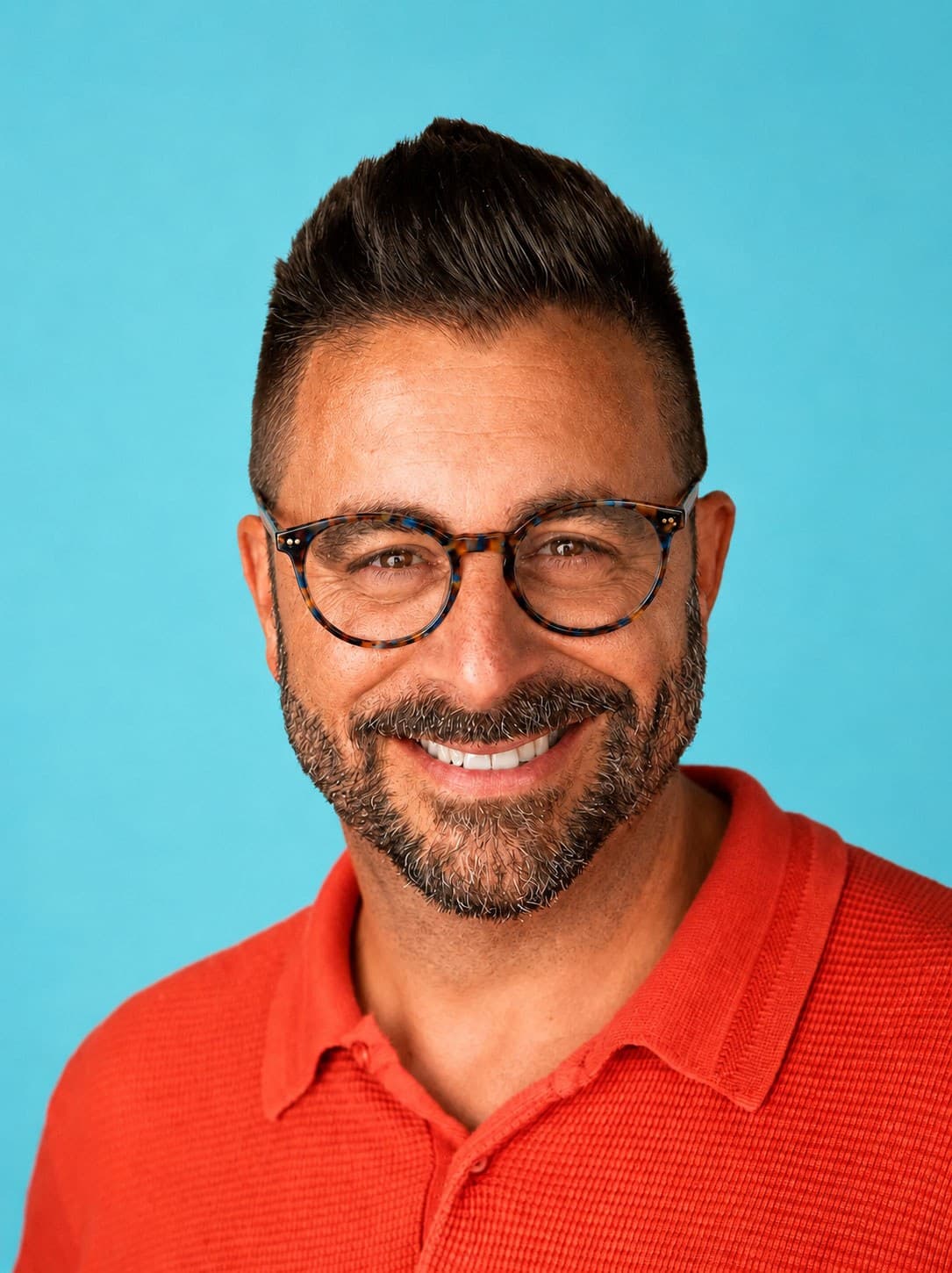 Smiling man with short dark hair, glasses, and a beard wearing an orange collared shirt, posing in front of a solid light blue background.
