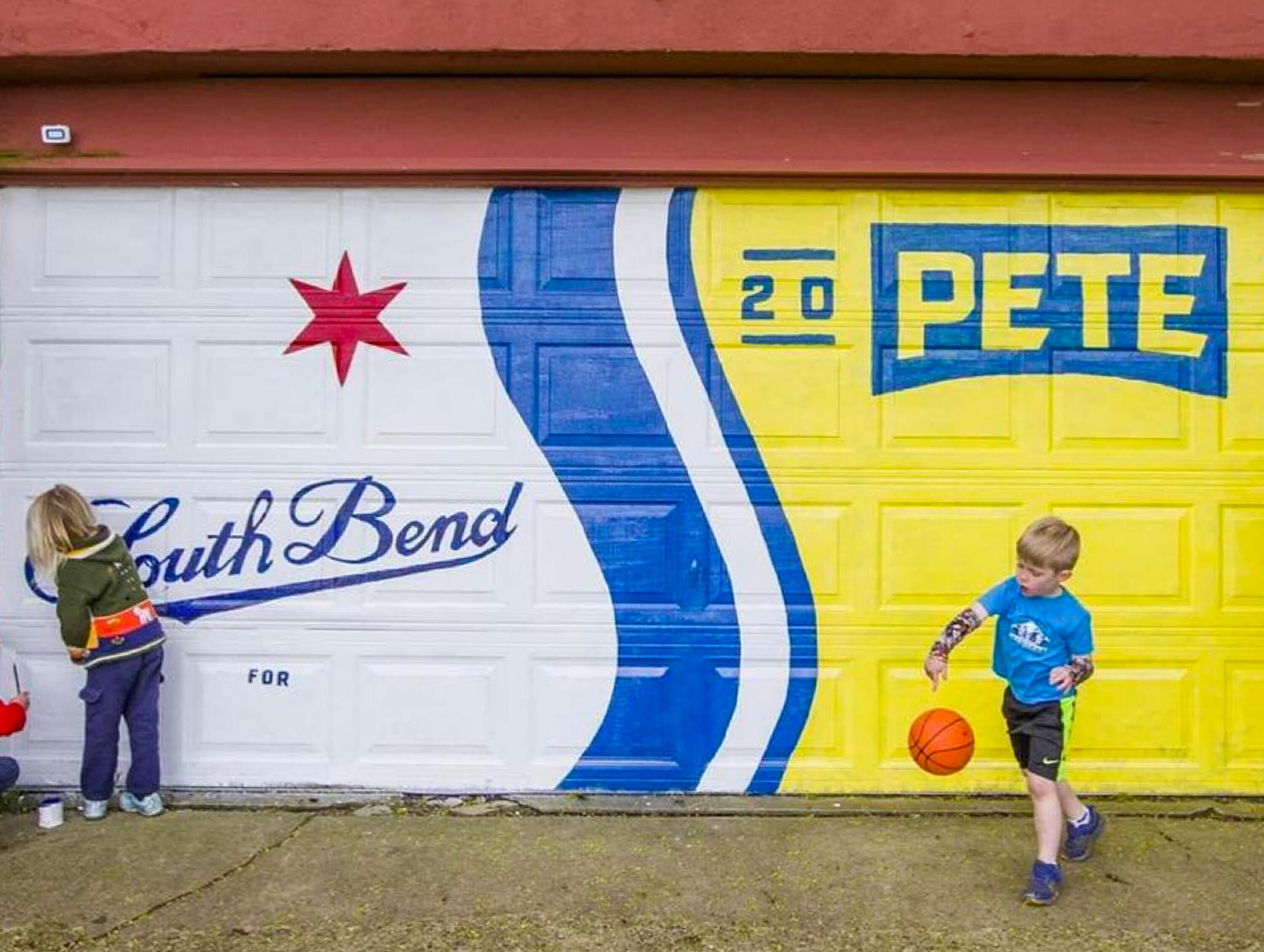 Two children play in front of a garage door painted with colorful artwork. The left side features the word "South Bend" and a red star, while the right side displays "20 PETE" on a yellow background. One child is painting, and the other is playing basketball.