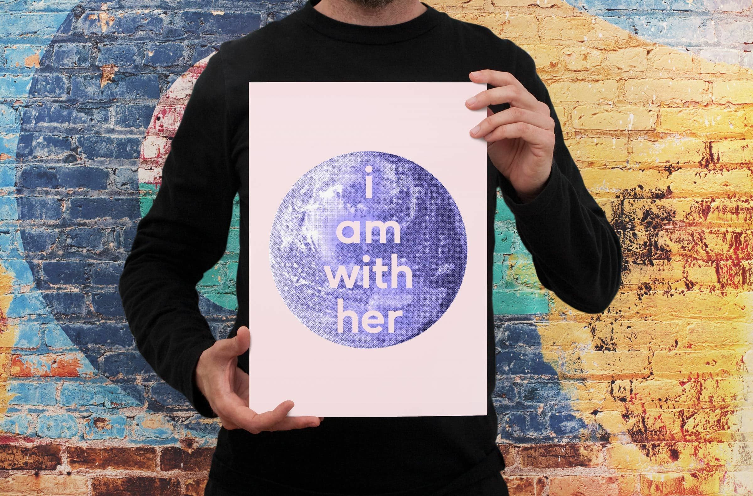 A person in a black shirt holds a poster featuring a blue and white globe illustration with the words "i am with her" on it. The background is a colorful, abstract mural painted on a brick wall.